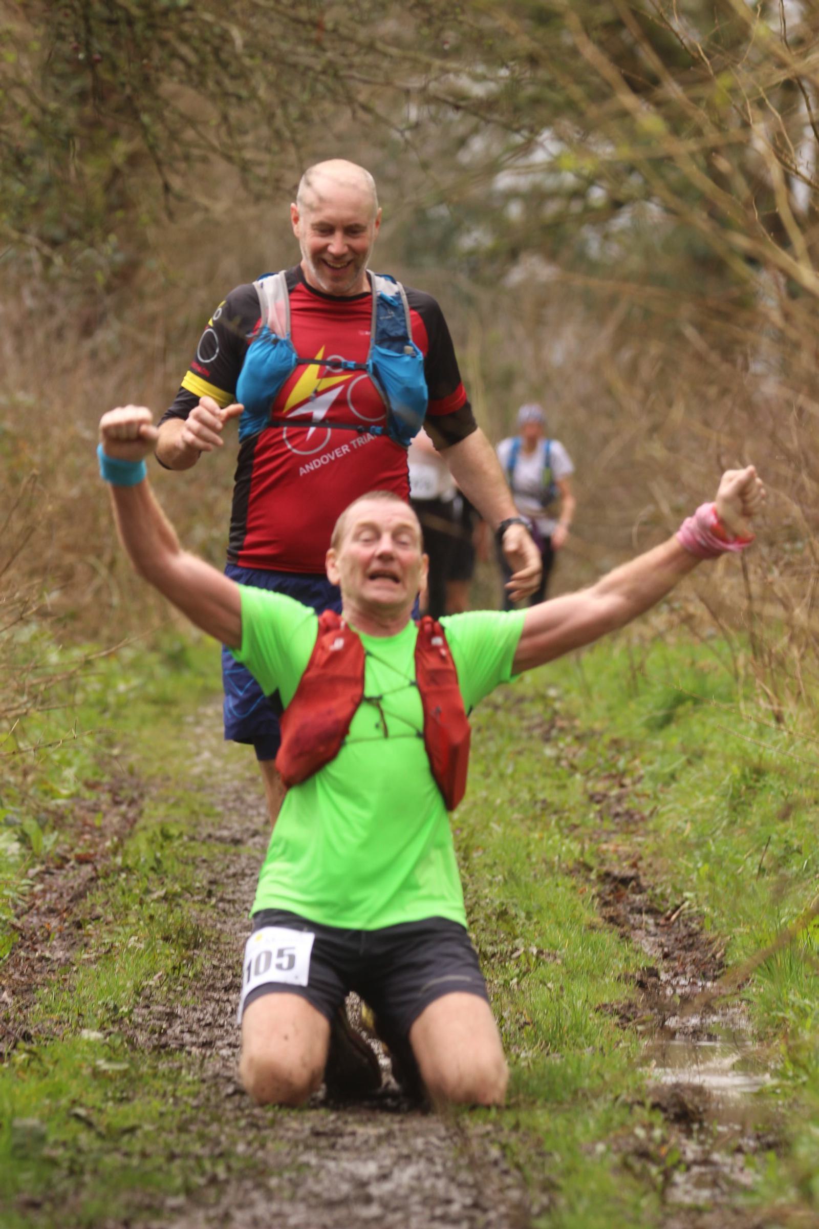 A runner in a green shirt and red vest kneels triumphantly in a muddy trail with arms raised. Another runner in a black and red shirt smiles behind him. Both appear to be participating in an outdoor race on a forested path.