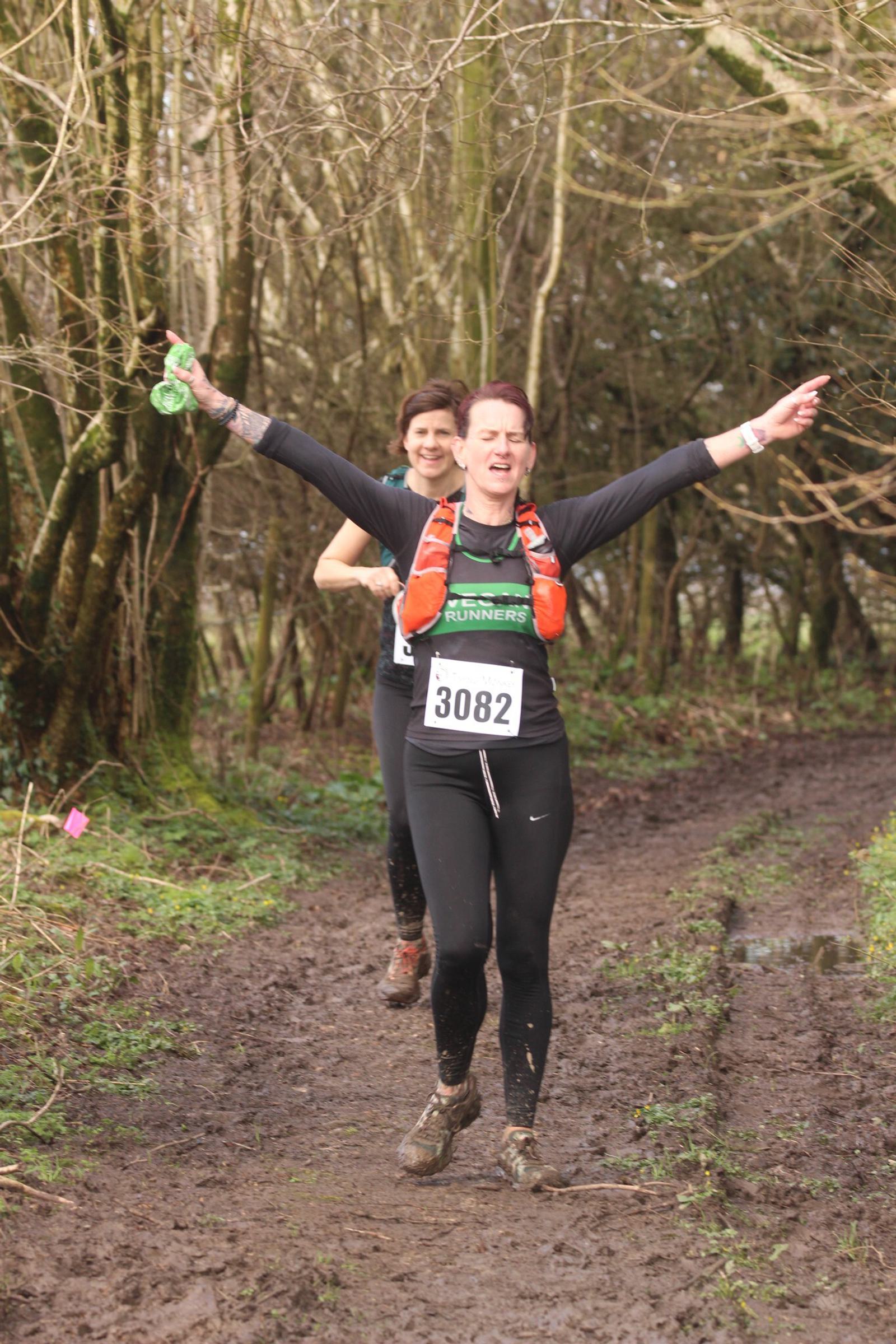 Two people running on a muddy forest trail. The person in front, wearing number 3082, smiles and raises their arms in celebration. Both wear black athletic clothing and hydration packs. Trees and pink markers line the path.