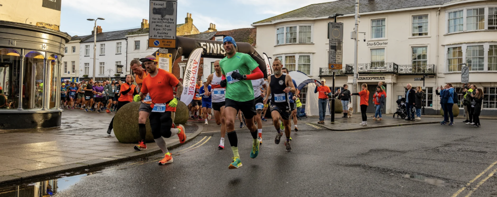 Runners in brightly colored athletic gear are participating in a street race, passing through a town with spectators lining the sidewalks. Various shops and buildings are visible in the background, along with a finish line banner further down the road.