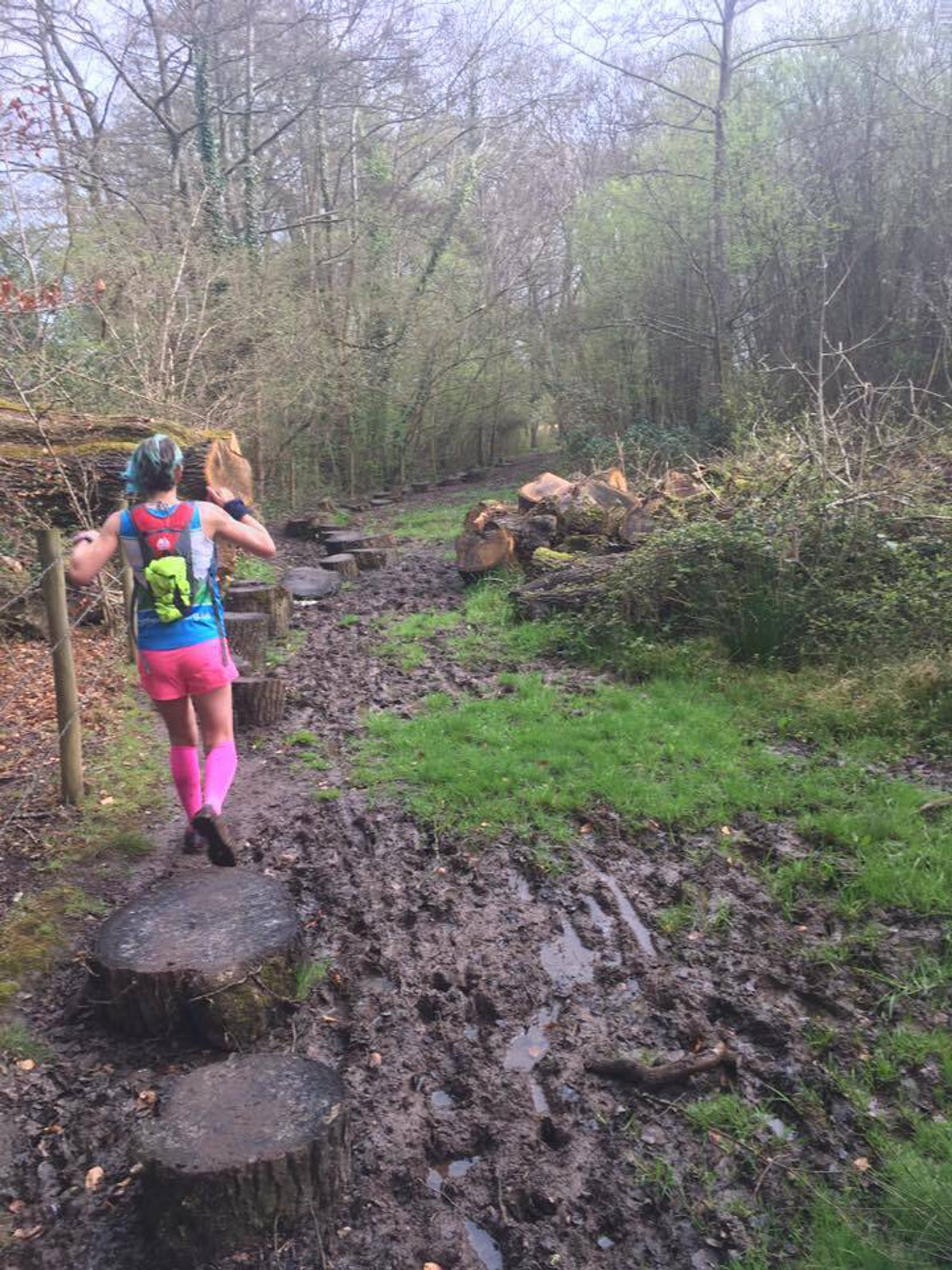 A person wearing a blue tank top, pink shorts, and pink knee-high socks is running on a muddy trail in a forest. They are balancing on large, round stepping stones made of tree stumps. The trees around them show sparse leaves, indicating early spring.
