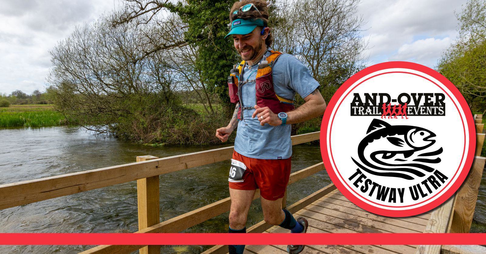 A runner in a blue shirt and red shorts crosses a wooden bridge during a trail event. He is wearing a headband and a hydration vest. The logo of "Andover Trail Events Testway Ultra" is displayed prominently. Trees and a river are in the background.