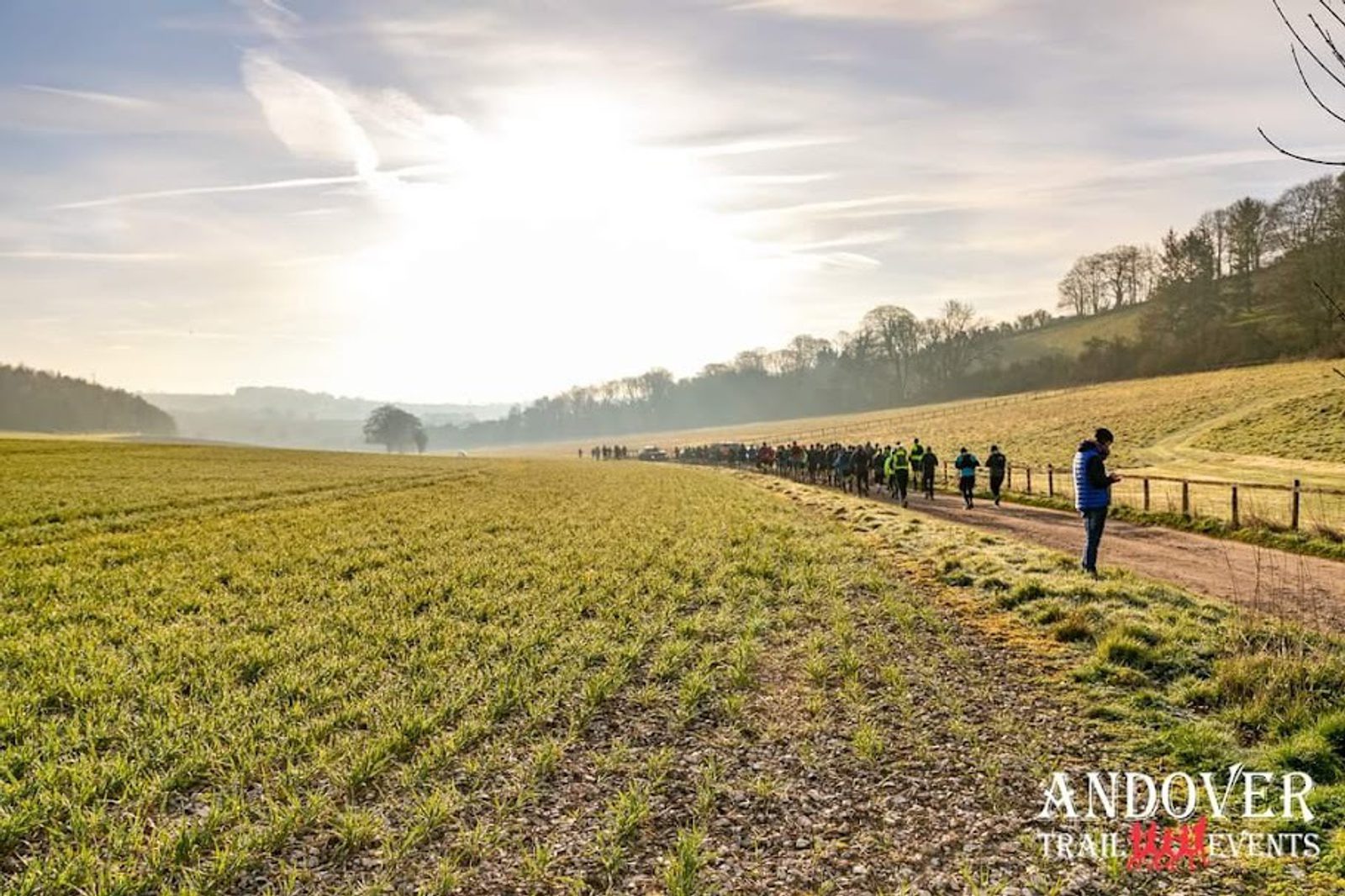 A line of runners on a trail winds through a sunlit, rural landscape with grassy fields and distant hills. A few trees dot the horizon. A man stands on the trail, possibly a race official. The logo "ANDOVER TRAIL EVENTS" is in the bottom right corner.