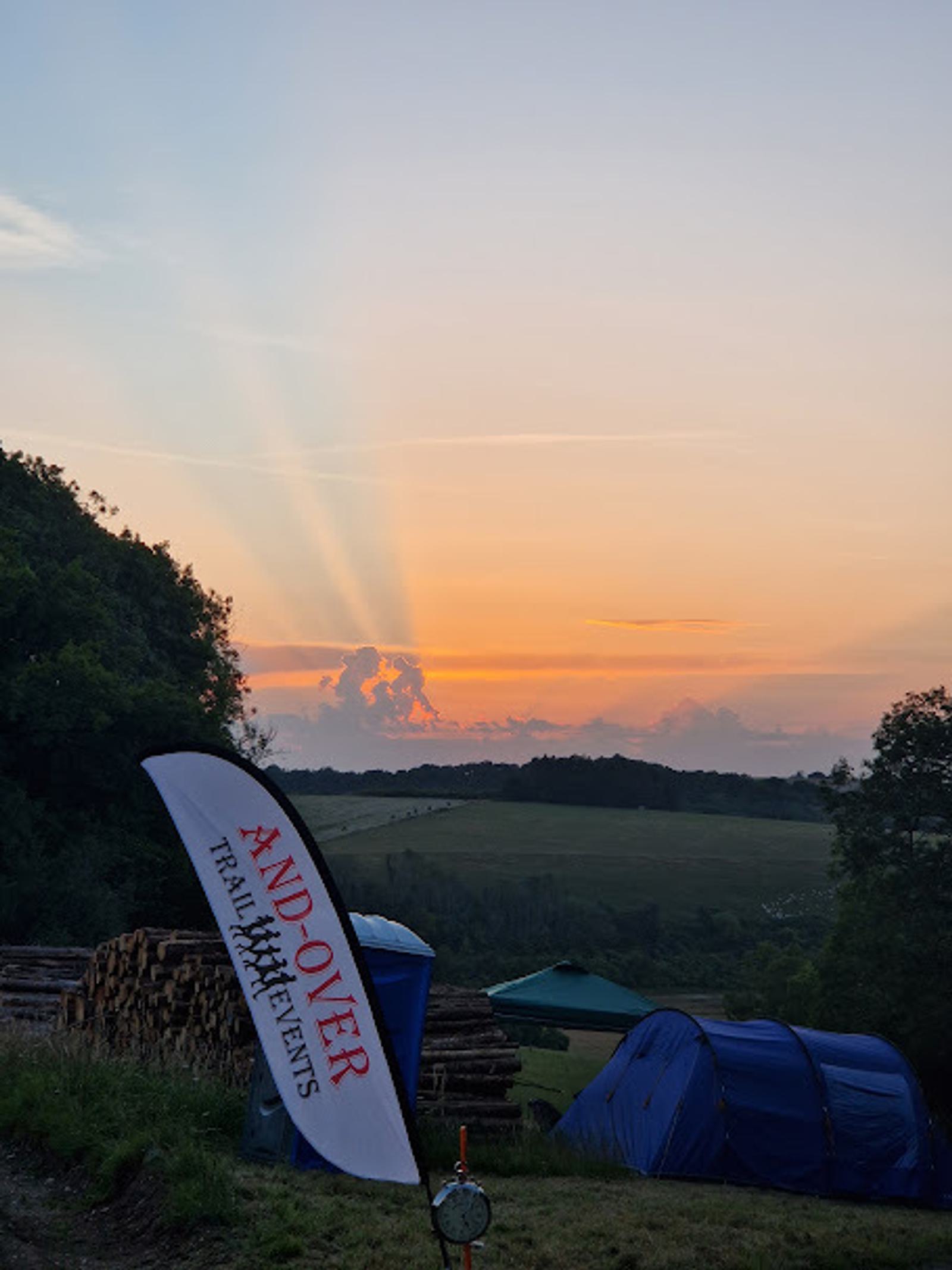 A scenic view of a sunset over rolling hills with a purple tent in the foreground. A flag reads "Andover Trail Events." Sun rays spread across the sky, and logs are stacked nearby.