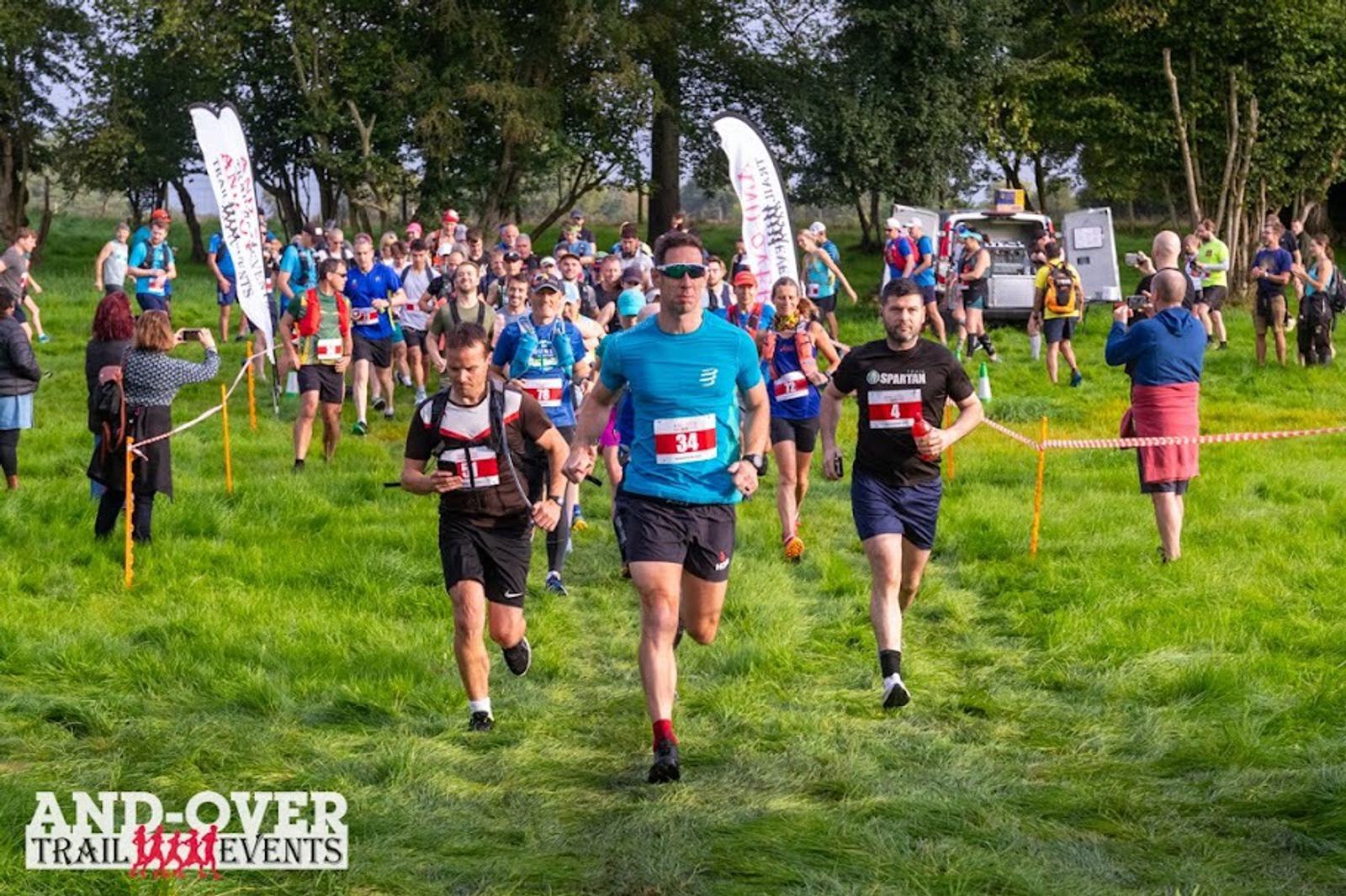 A group of runners participates in an outdoor trail event, moving through a grassy field. They wear numbered bibs, and some spectators can be seen in the background. Banners and trees line the course.