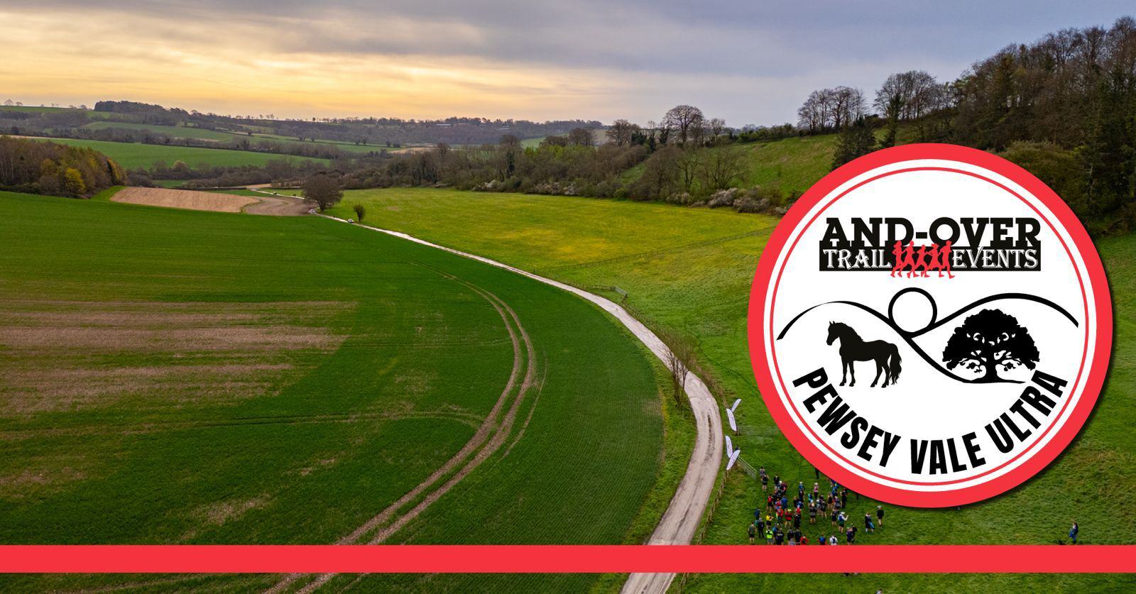 A scenic trail with green fields and distant hills under a cloudy sky. A winding path with small groups of people is visible. In the foreground, a large logo for the Andover Trail Events Pewsey Vale Ultra is present.