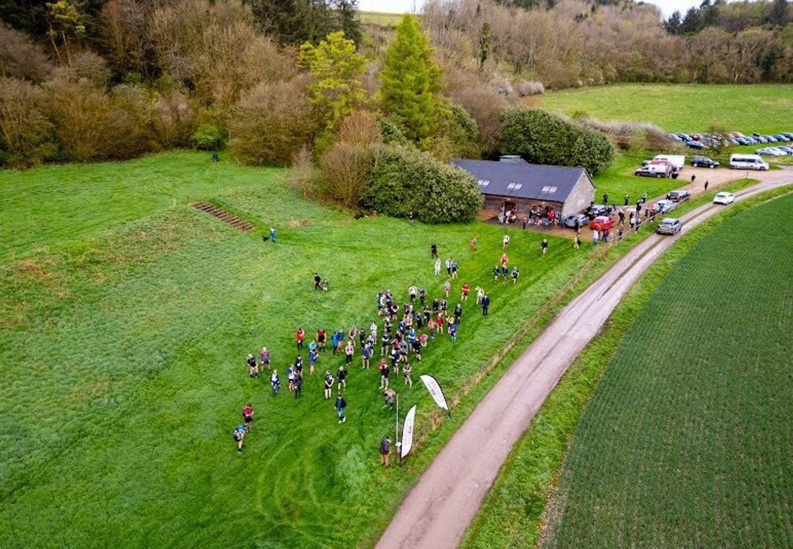 Aerial view of a trail event starting line. Participants are gathered on a grassy field near a large barn-like building. A winding dirt road and a row of parked cars are visible. Trees and open fields surround the area. "ANDOVER TRAIL EVENTS" logo is in the bottom left corner.