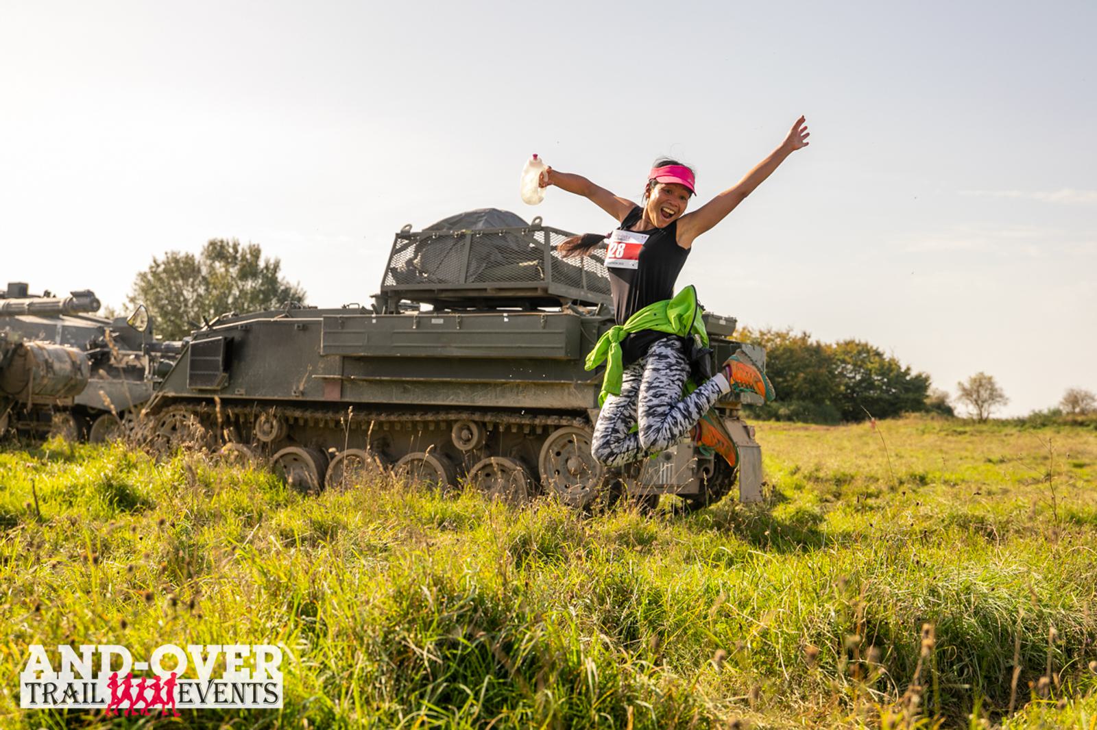 A person joyfully jumps in front of a parked tank on a grassy field during a trail event. They are wearing a pink headband, black top, and patterned leggings. The sky is clear and sunny, with a logo saying "Andover Trail Events" in the corner.