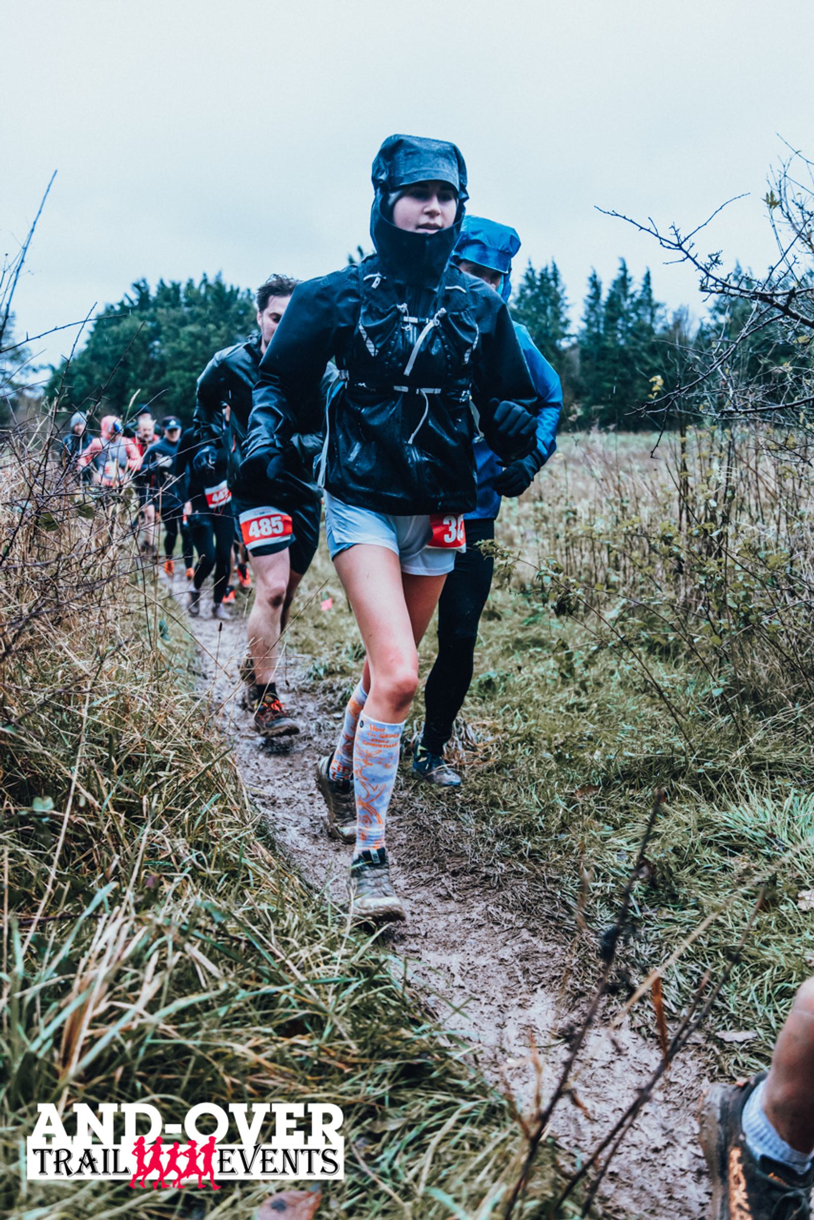 Participants in trail running gear jog along a muddy path during an outdoor race, surrounded by bushes and trees. The sky is overcast, and a logo at the bottom reads "Andover Trail Events.