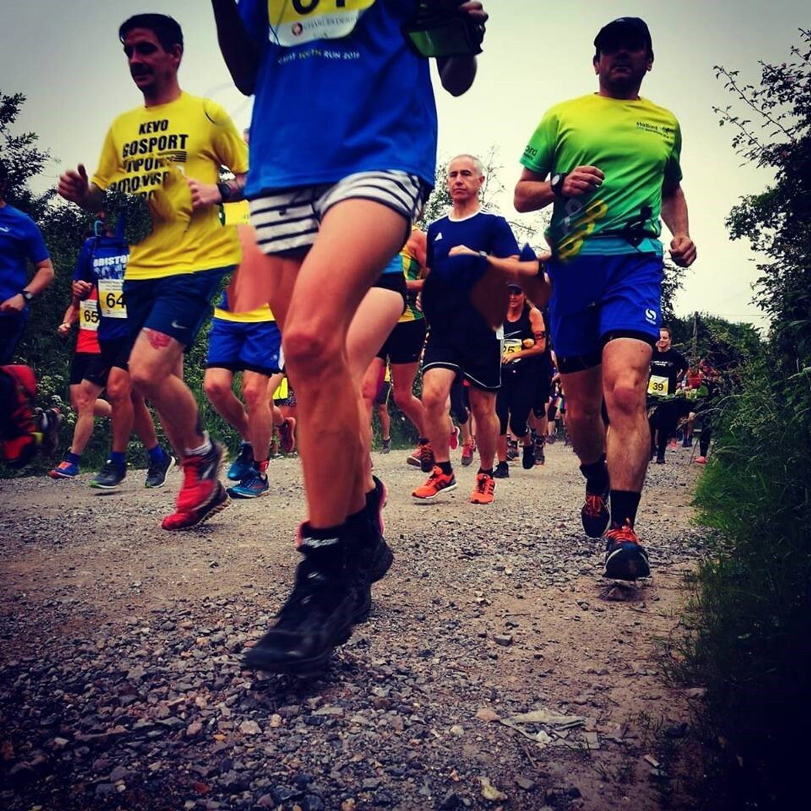Runners in colorful athletic gear compete on a gravel path during an outdoor race. The scene captures movement and energy, with trees and greenery lining the background.