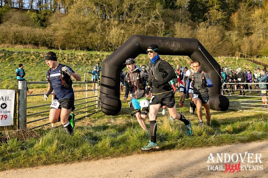 Runners participating in the Andover Trail Events race. They are running on a path surrounded by greenery and trees, passing under a large black inflatable arch. The weather appears clear and sunny.