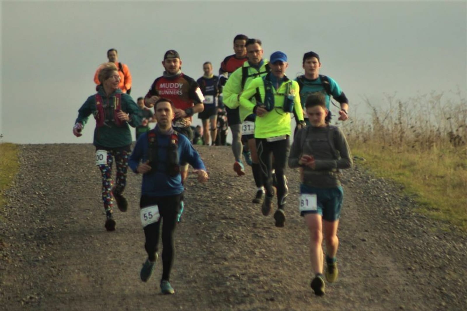 A group of runners is participating in a trail race on a gravel path. Some wear colorful running gear, and several have race bibs pinned to their clothing. The background is cloudy, and the surrounding landscape is grassy. The runners appear to be focused and determined.