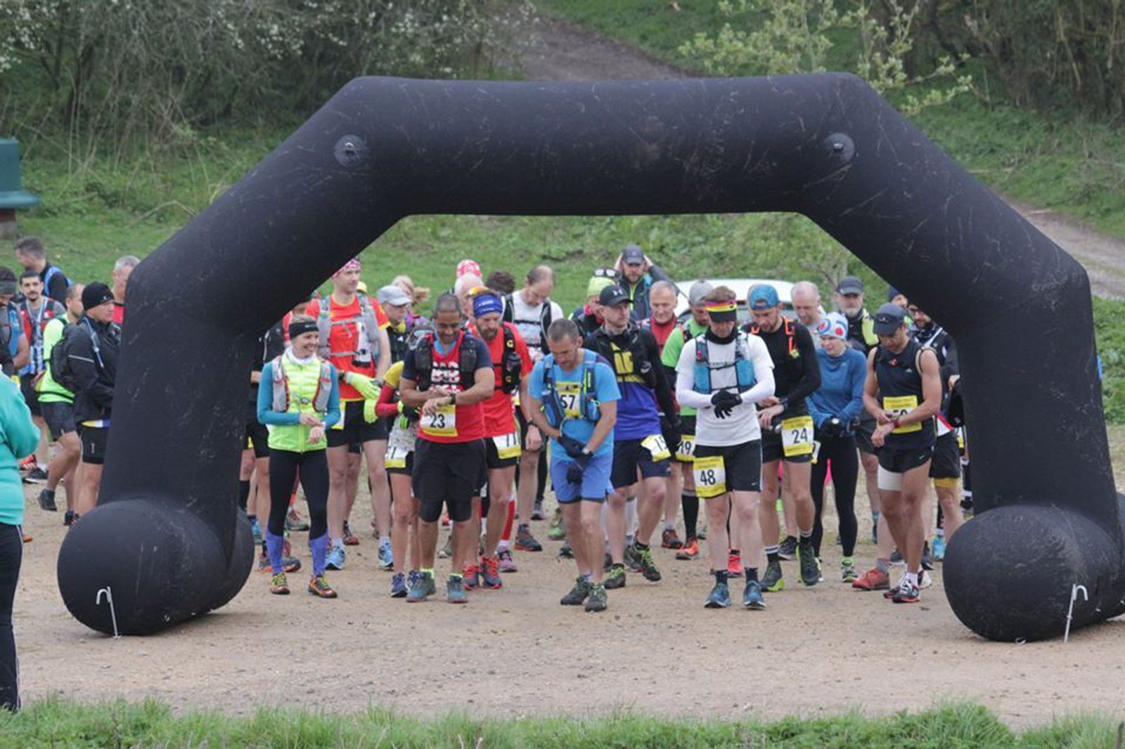 A group of runners gathered at the starting line under a large black inflatable arch, preparing to begin a race. They are outdoors on a gravel path, surrounded by greenery. The participants are wearing various running gear and race bibs with numbers.