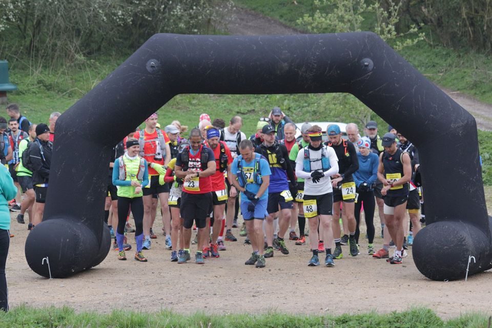 A group of runners gathered at the starting line under a large black inflatable arch, preparing to begin a race. They are outdoors on a gravel path, surrounded by greenery. The participants are wearing various running gear and race bibs with numbers.