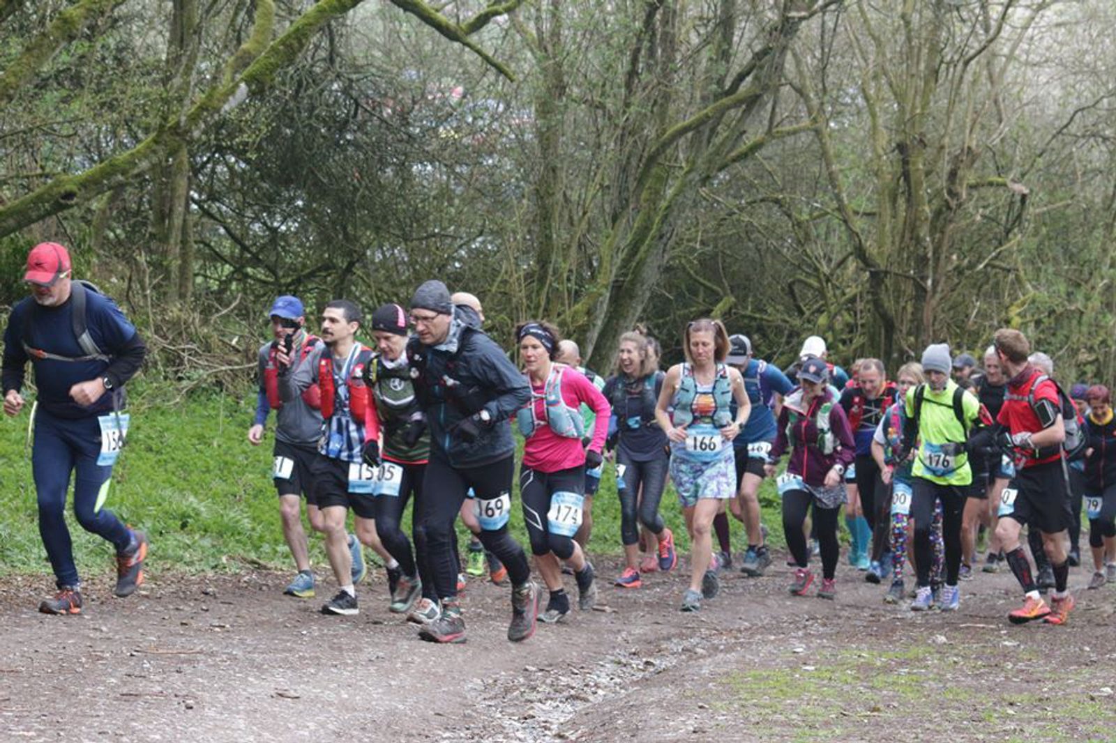 A group of people wearing running gear, numbered bibs, and hydration packs run along a dirt path in a lush forest. The runners navigate a slight uphill section surrounded by trees and greenery on both sides. The atmosphere appears energetic and focused.