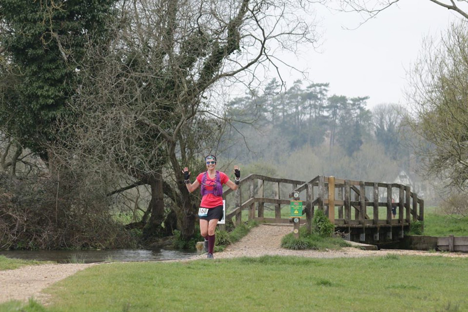 A runner in a pink shirt, black shorts, and maroon socks with number 159 on their shorts is running on a gravel path near a small wooden bridge. Trees and greenery surround the area, and the runner is raising both arms in a cheerful gesture.
