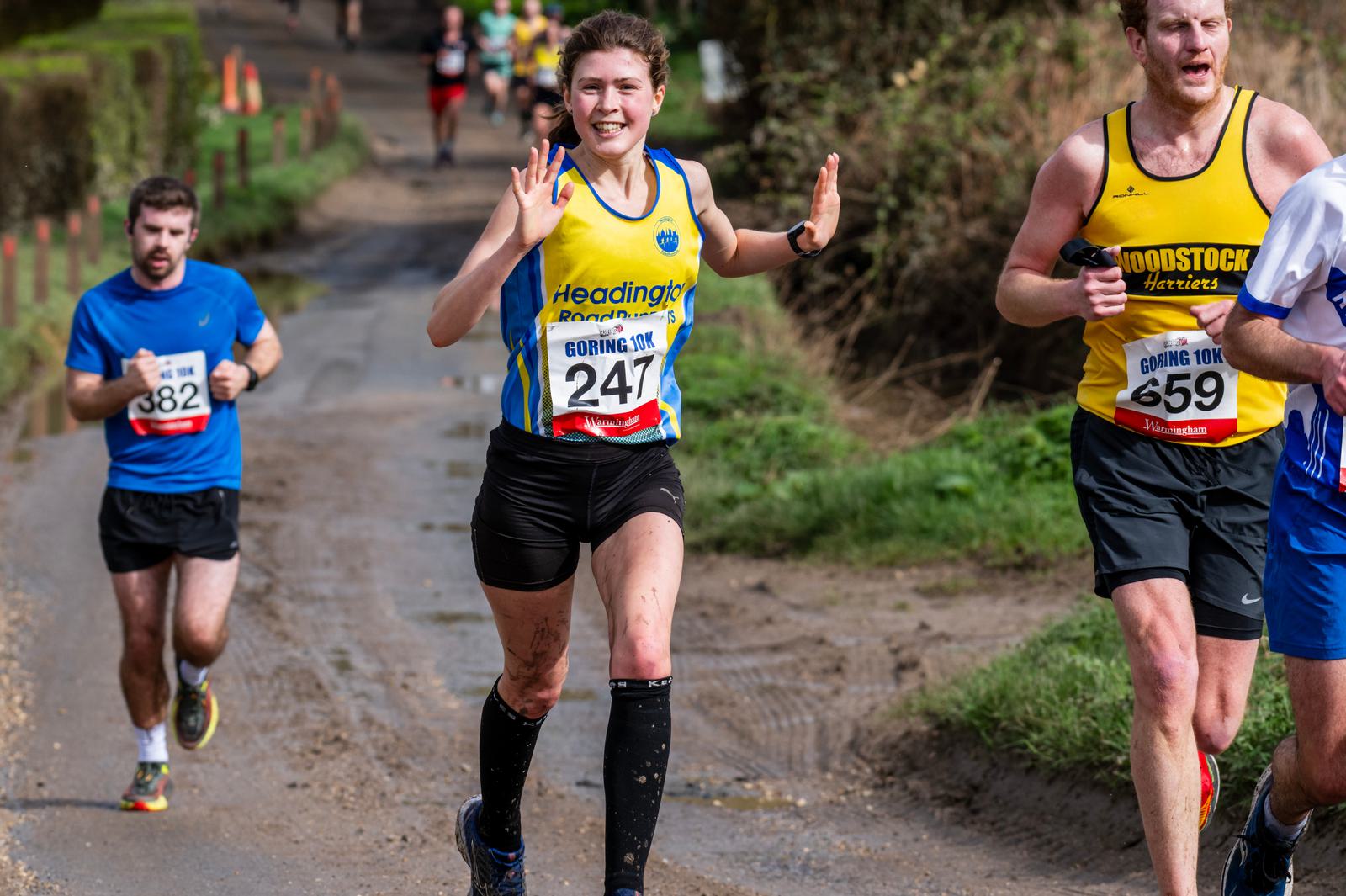 A woman in a blue and yellow running outfit, wearing the number 247, smiles and waves at the camera while running a race. Two other runners are visible nearby on a muddy path. The scene is outdoors with greenery in the background.