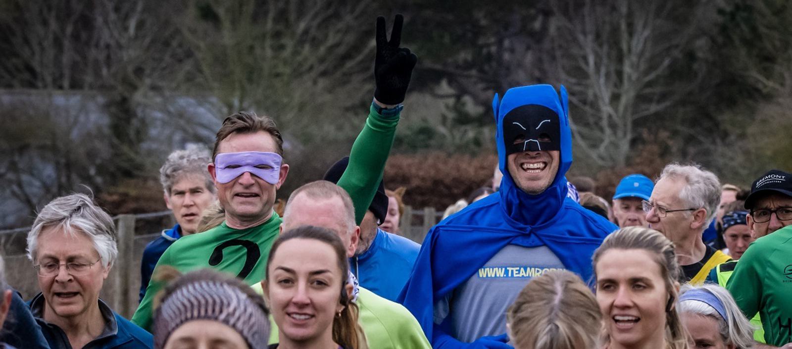A group of people running in an outdoor event, with two individuals wearing superhero costumes. One is dressed in a black and blue cape, and the other in a green suit with a purple mask. The atmosphere is lively and cheerful.