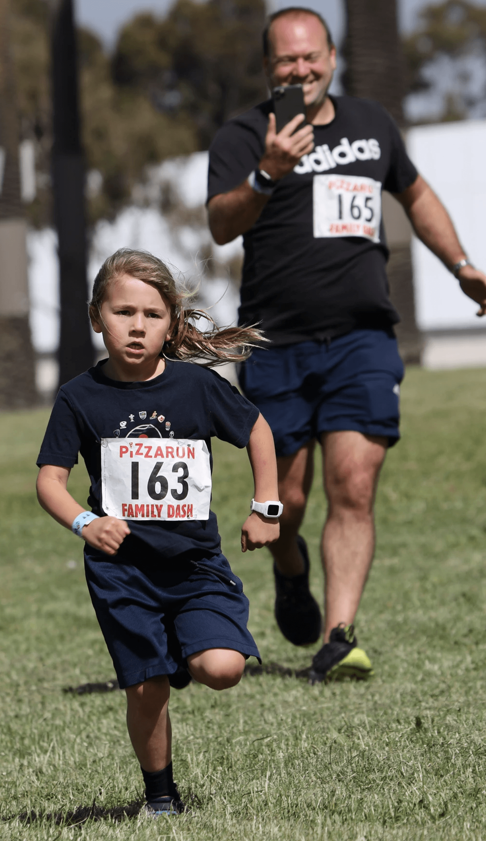A young child with long hair, wearing a navy blue outfit and a race number "163", runs on a grassy field with an older person, also in athletic attire, and race number "165" running behind. They participate in the "PIZZARUN FAMILY DASH.