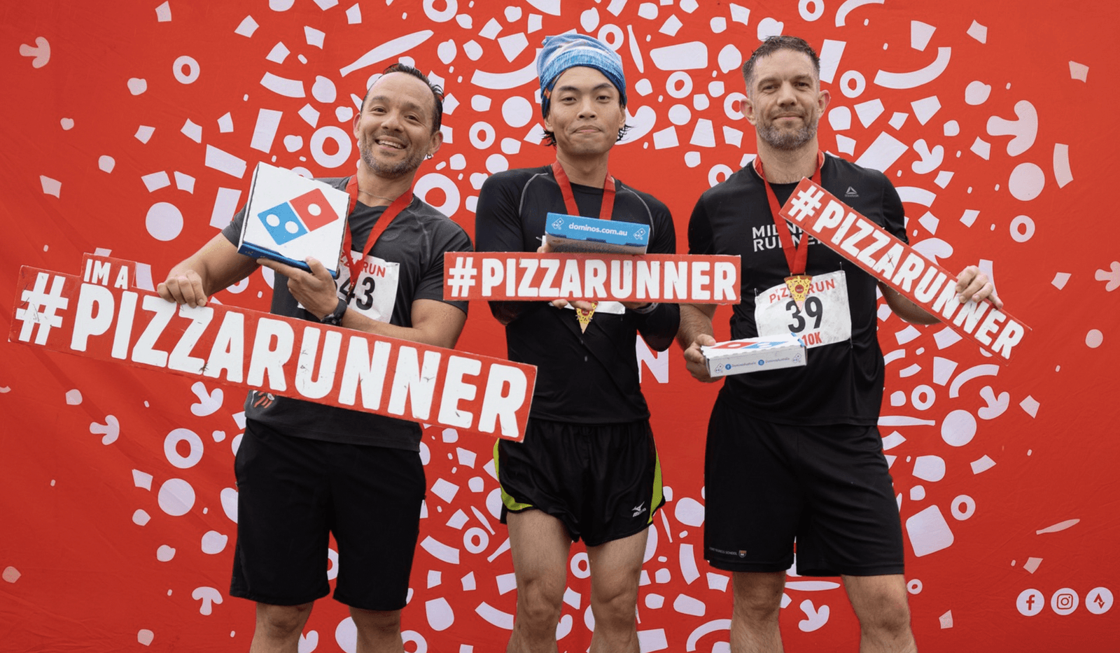 Three smiling men in athletic gear hold pizza boxes and large hashtags that read #PIZZARUNNER. They stand in front of a red backdrop covered with pizza-themed graphics. Each wears a race bib, indicating participation in a themed fun run event.
