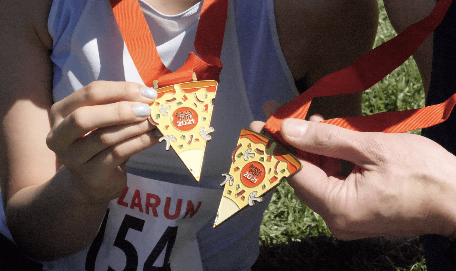 Two individuals display pizza slice-shaped medals with red ribbons around their necks. The medals are engraved with "Pizza Run 2021." The partial view of one person's race bib shows the number 154. The background is grassy, suggesting an outdoor event.
