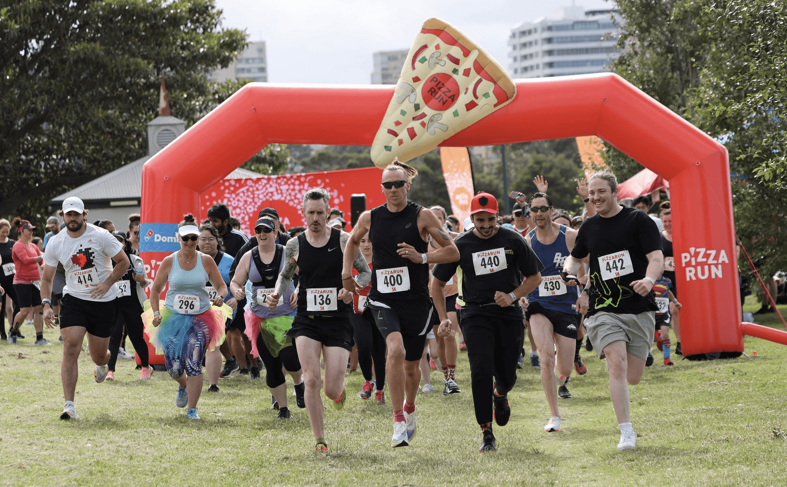A group of runners wearing bibs are starting a race under an inflatable arch that reads "Pizza Run." One runner is dressed in a large pizza slice costume. Trees and buildings are visible in the background. The atmosphere is lively with participants of various ages.