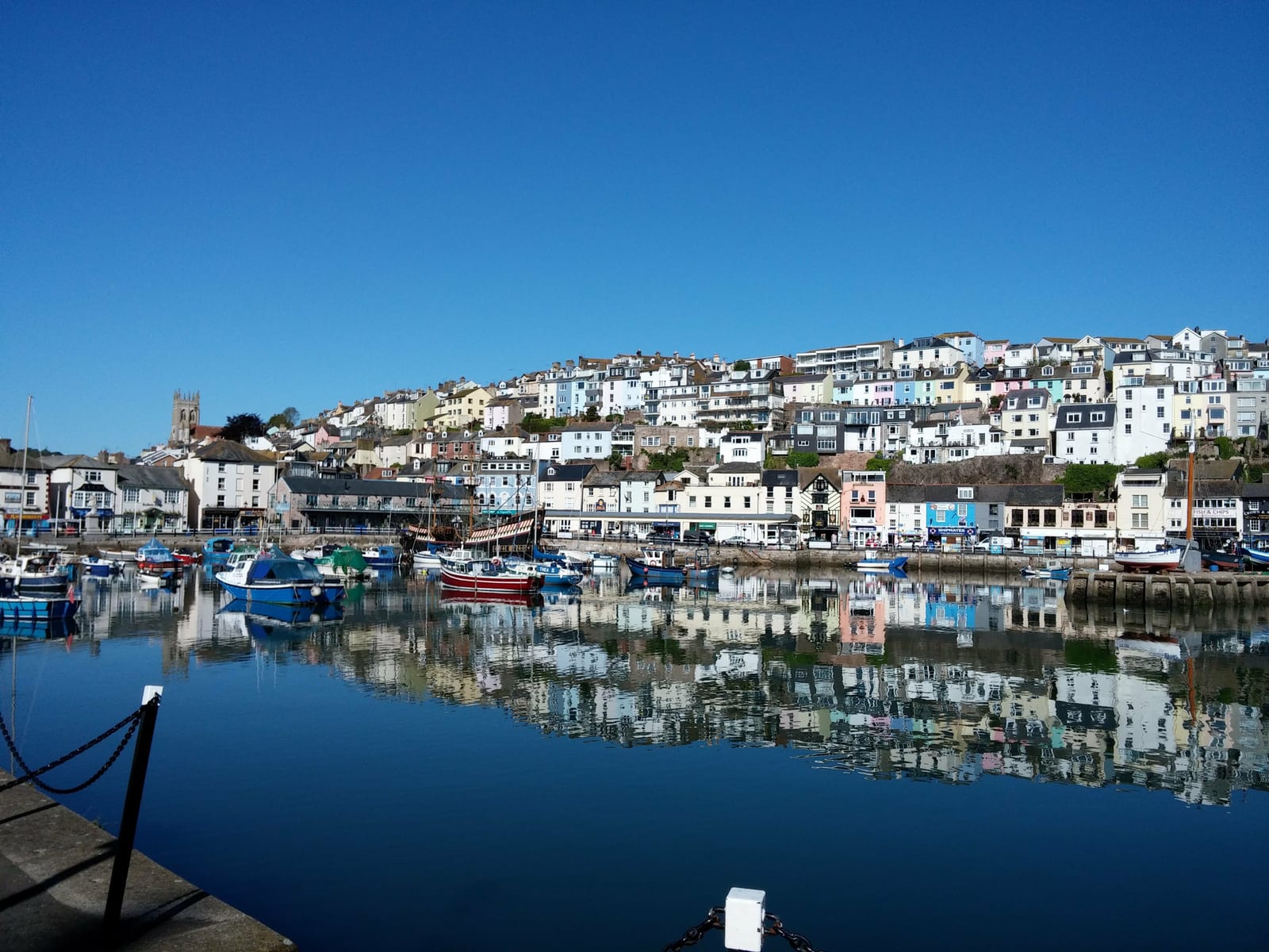 A picturesque coastal town with colorful houses stacked on a hillside, reflected in the calm waters of a marina. Various boats are docked in the harbor, and the sky is clear and blue. A church tower is visible among the houses.