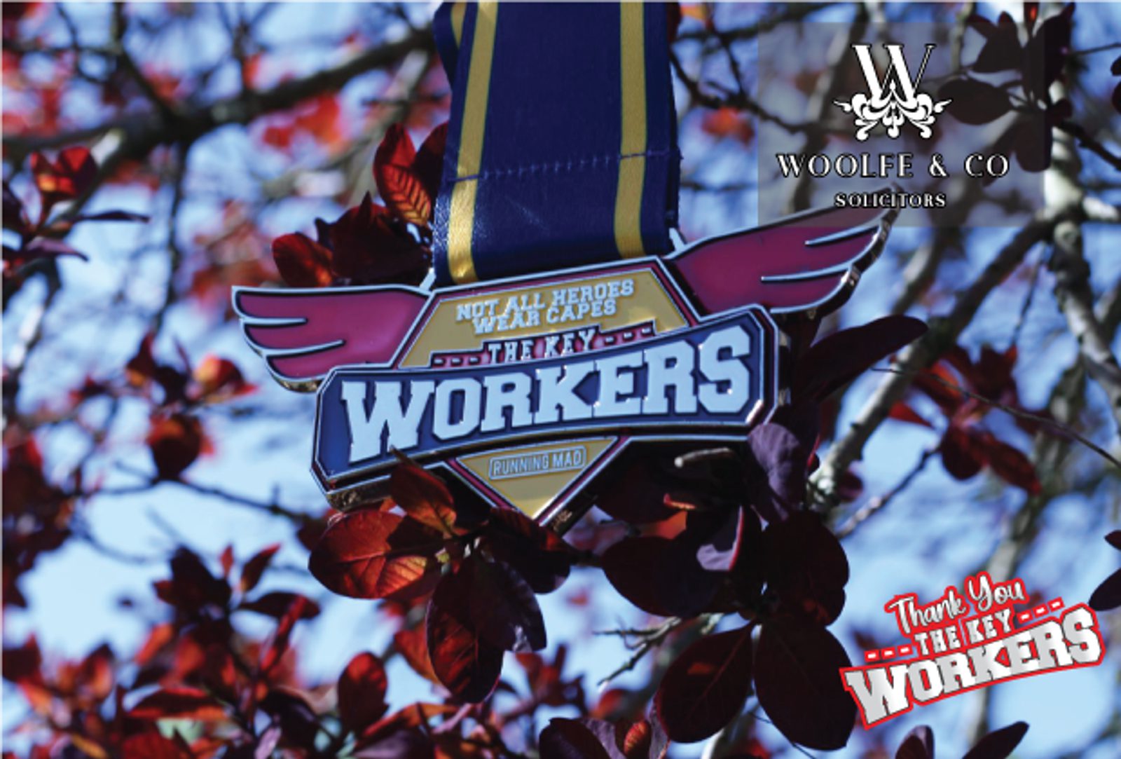 An image of a "Thank You Key Workers" medal hanging from a tree branch with red leaves. The medal's ribbon is blue and yellow, and text on the medal reads "Not All Heroes Wear Capes" and "Thank You Key Workers." Logos for "Woolfe & Co Solicitors" and "Thank You The Key Workers.co.uk" are visible.