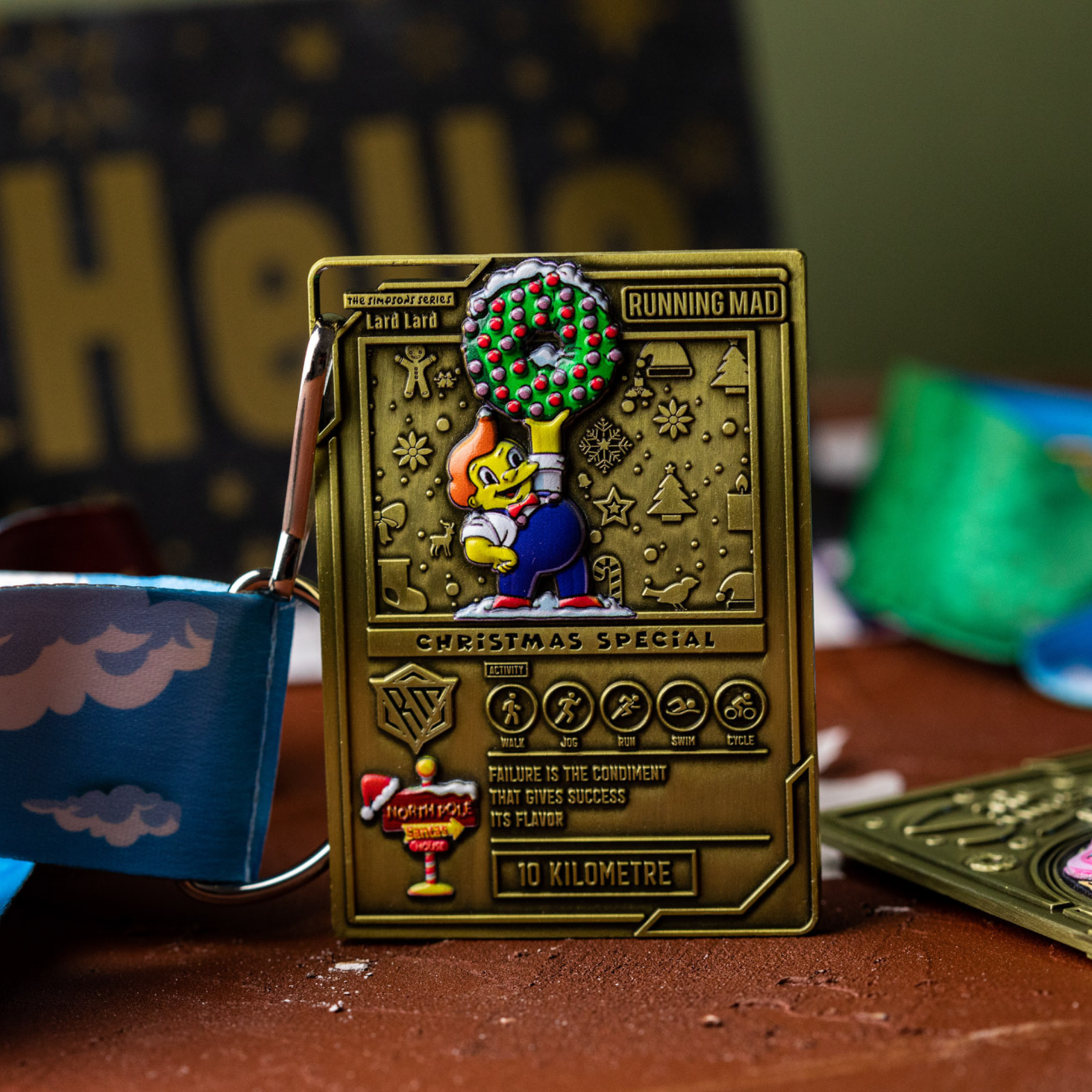 A golden Christmas-themed medal with a cartoon character holding a decorated tree. Text reads "Running Mad Christmas Special" and "10 Kilometre." It's attached to a blue and white lanyard on a table with colorful items in the background.