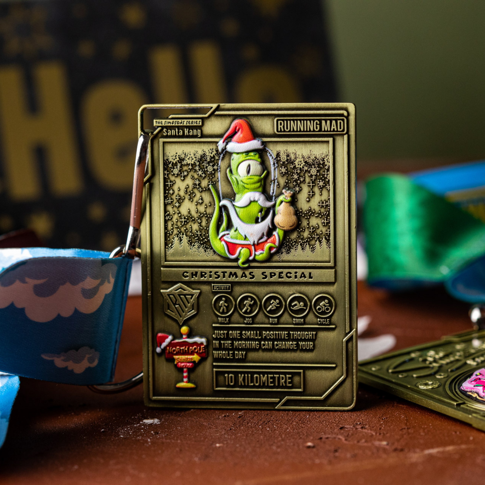 A gold medal featuring a green character in a Santa hat surrounded by snowflakes. Text reads "Christmas Special" and "10 Kilometre." A blue and white ribbon is attached. Background shows another medal partially visible.