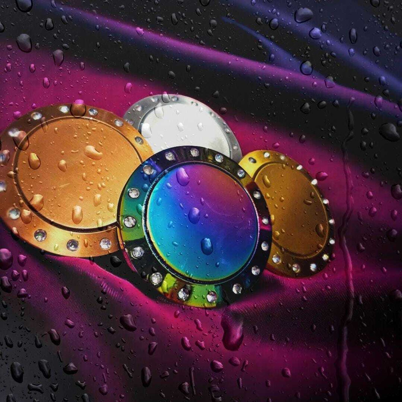 Colorful metallic rings with rhinestones rest on a vibrant purple and black background, covered in water droplets. The rings are in various shades, including gold, silver, and multicolored iridescent.