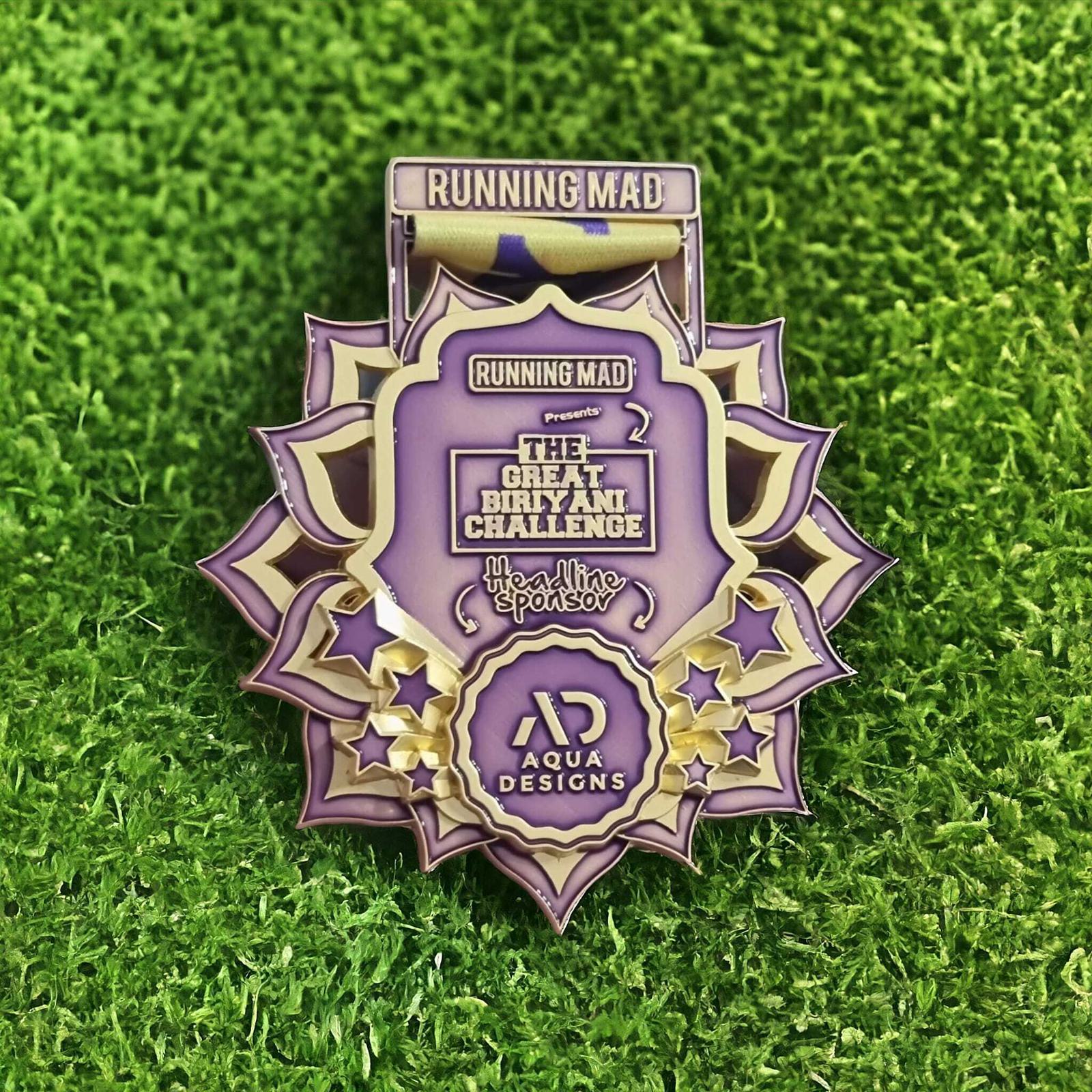 A decorative purple and gold medal on a grassy background, featuring text that reads "Running Mad presents The Great Biryani Challenge" with "Headline Sponsor" and "Aqua Designs" logos displayed. The medal includes star patterns.