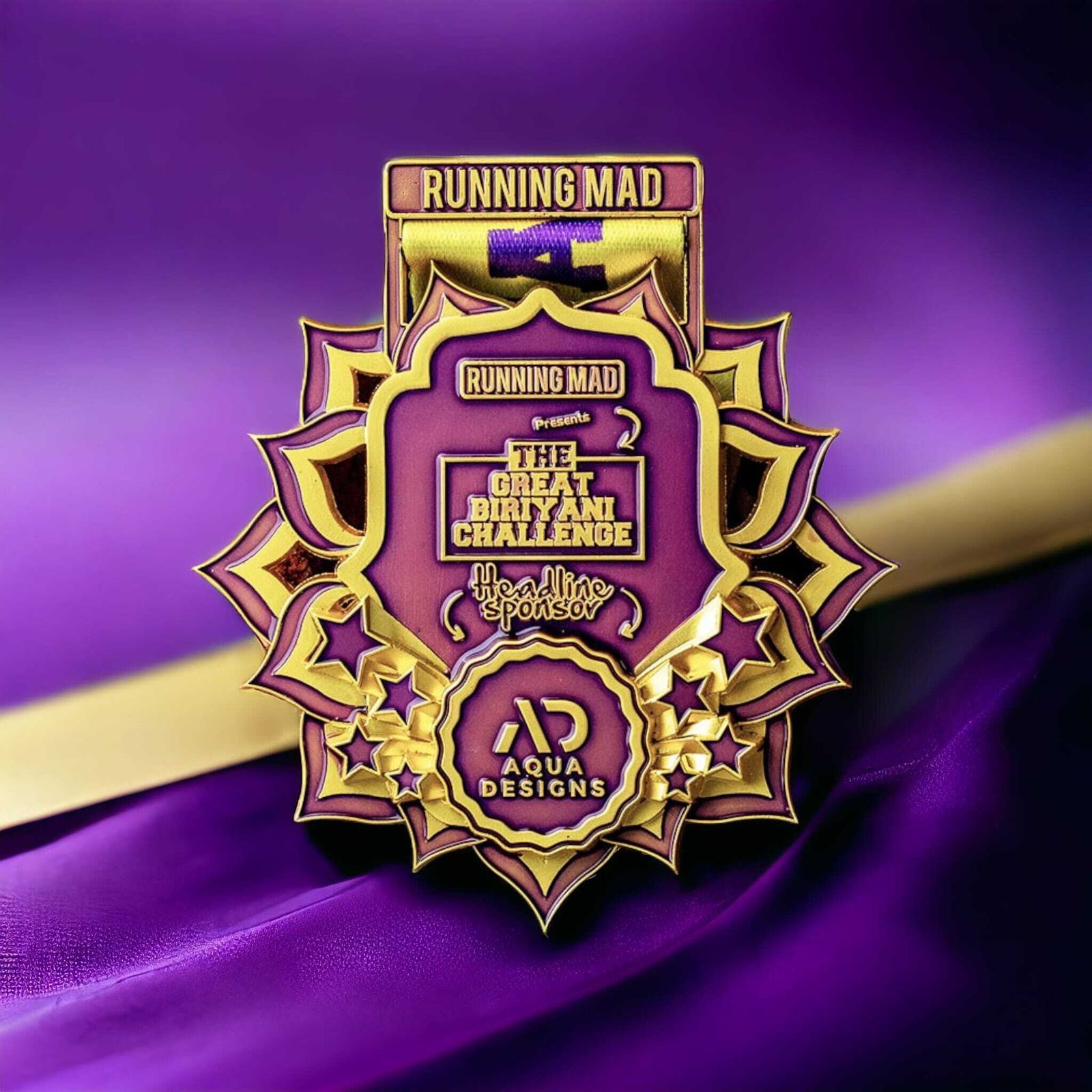 A vibrant badge featuring a gold and purple design. The text reads "Running Mad presents The Great Biryani Challenge" with star decorations and "Aqua Designs" at the bottom. The background is a rich purple fabric.