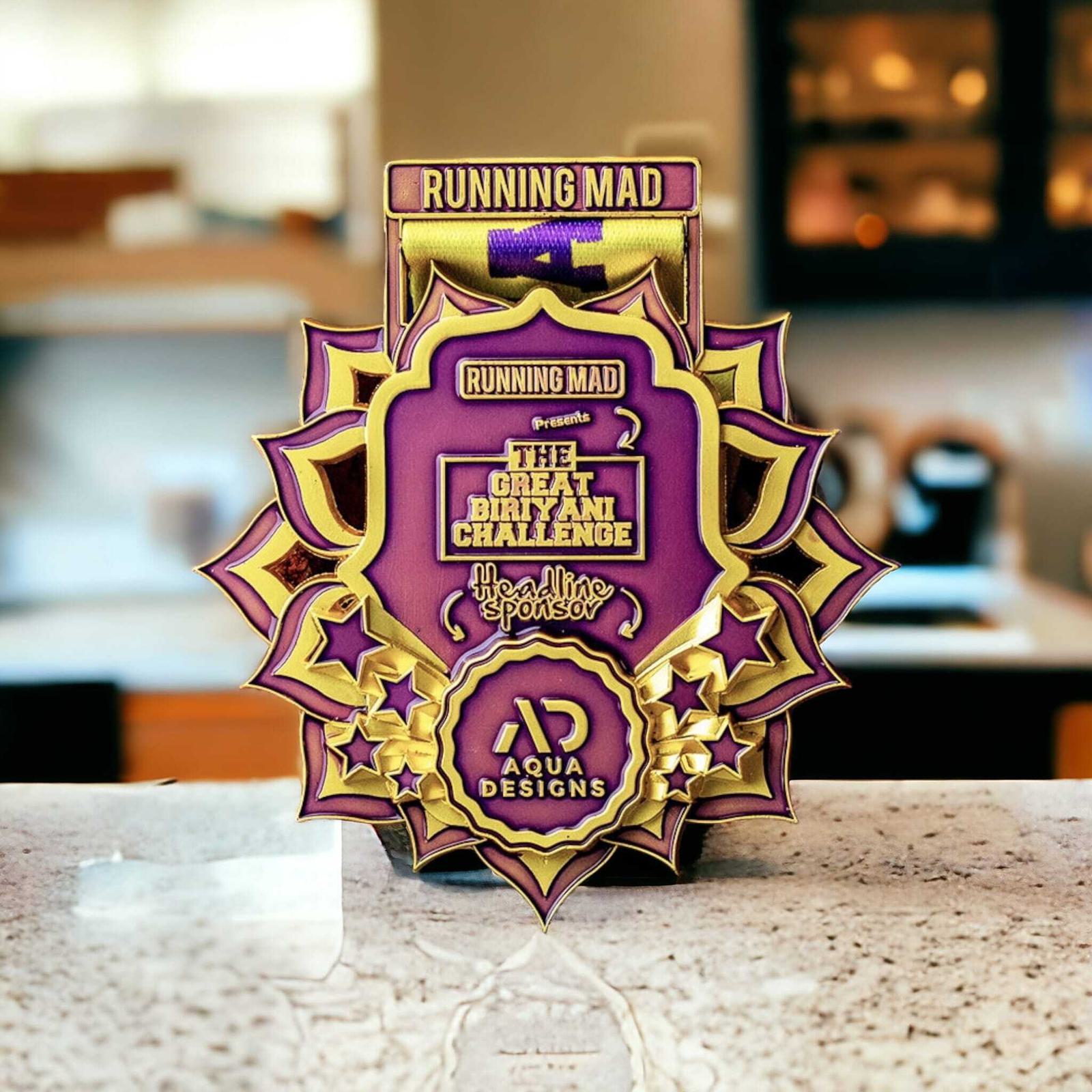 A gold and purple medal with the text "Running Mad presents The Great Biriyani Challenge" is displayed on a kitchen countertop. The sponsor, Aqua Designs, is mentioned at the bottom of the medal.
