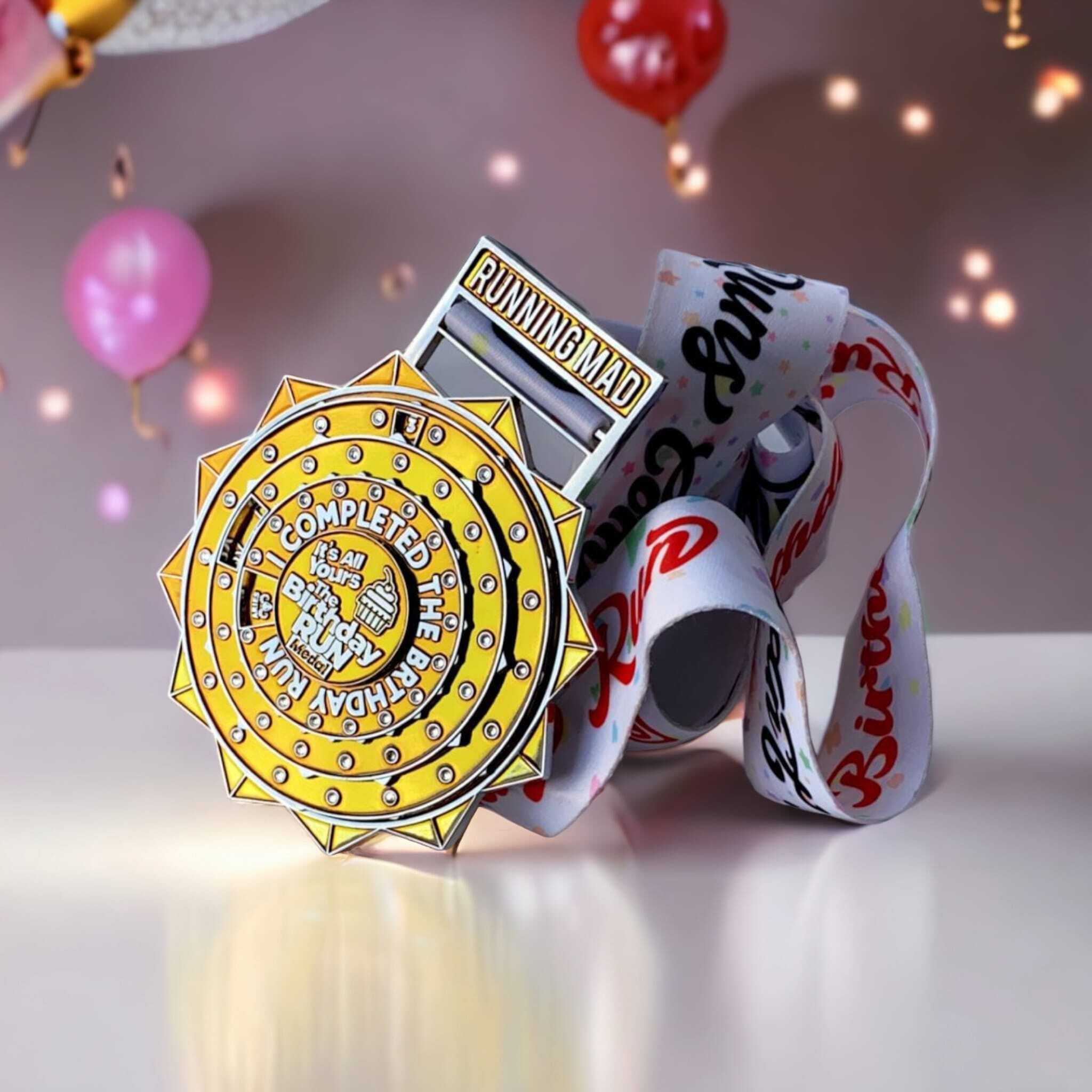 A colorful medal with a yellow sunburst design, labeled "Running Mad" and "I Completed The Birthday Race," rests on a table. The attached white ribbon is decorated with various colors. Blurred balloons are visible in the background.