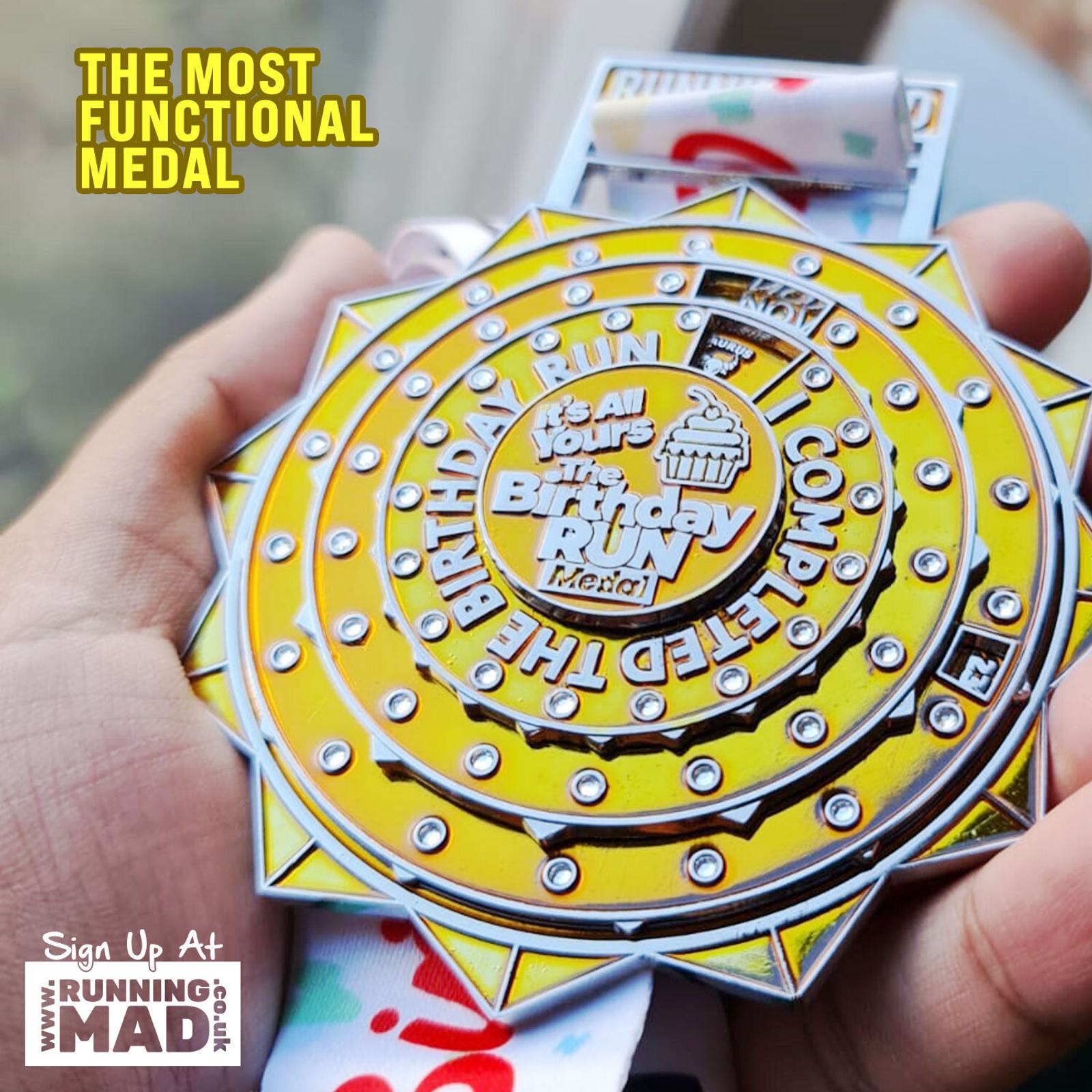 A hand holds a brightly colored birthday-themed running medal with text "THE MOST FUNCTIONAL MEDAL" and "Birthday Run Complete." The medal is intricately designed with a cupcake image. Website text visible: "Sign Up At RUNNINGMAD.CO.UK.