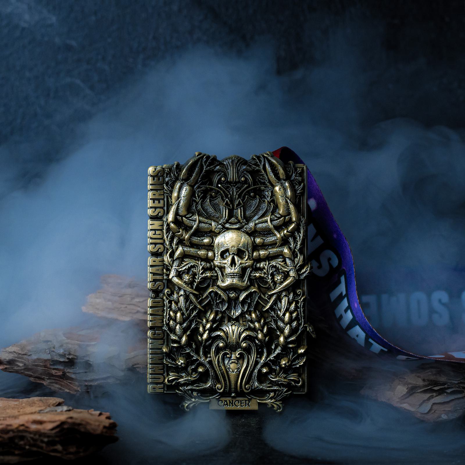 A dark, intricate medal with a skull design and the word "CANCER" at the bottom, surrounded by smoky effects. The medal is suspended by a ribbon and rests against a textured background with wooden elements.
