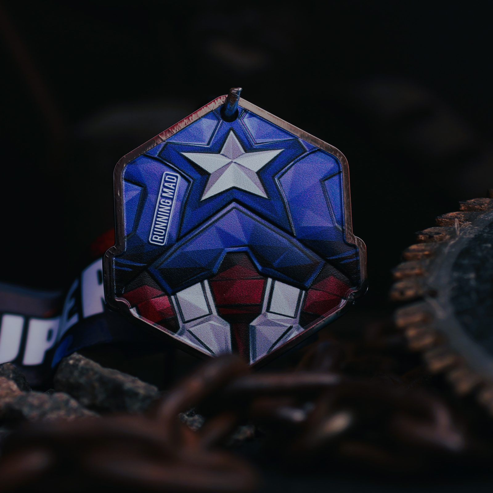 A hexagonal medal with a superhero emblem featuring a white star on a blue background, red and white stripes, and "RUNNING MAD" text. It hangs on a lanyard among metal chains and gears against a dark background.
