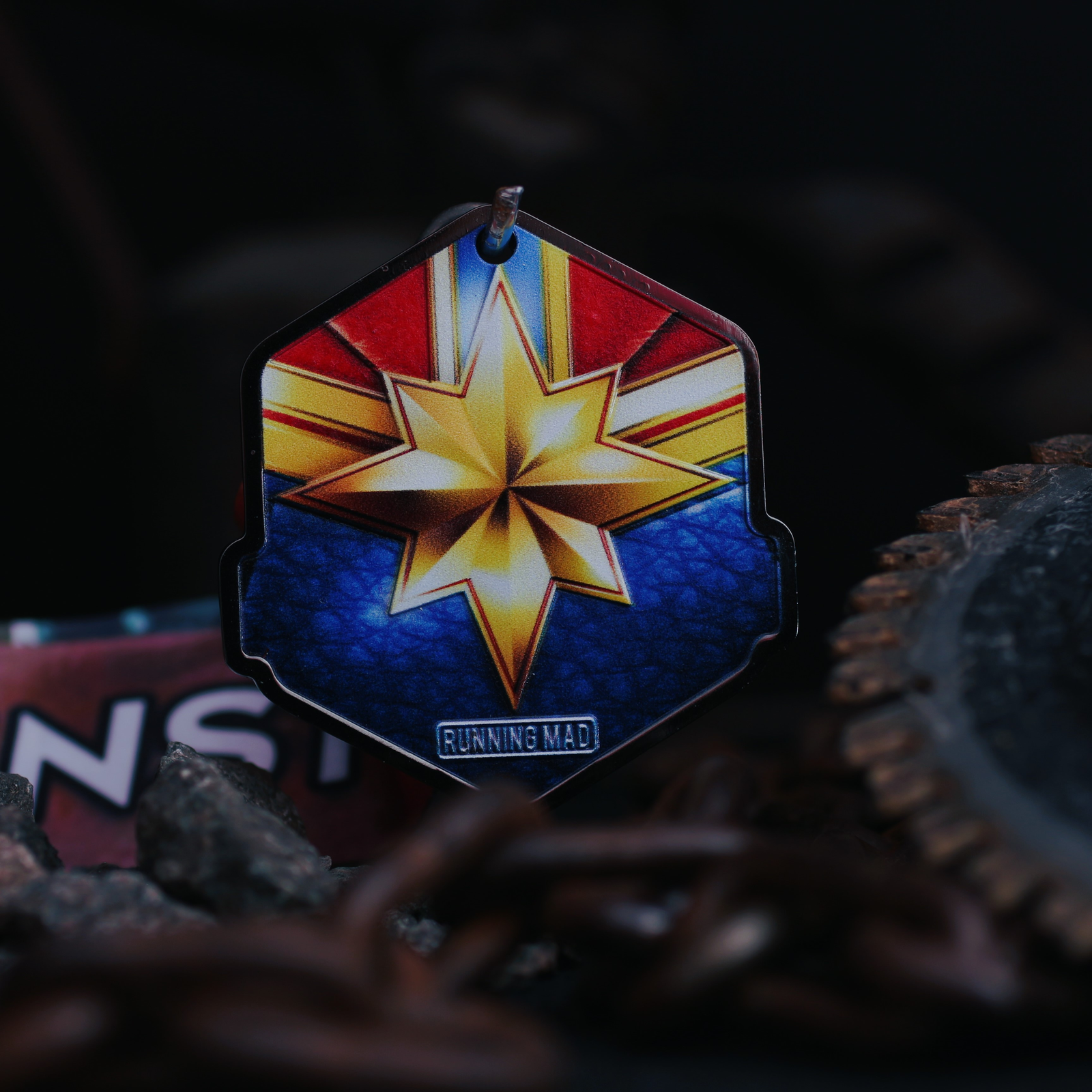 A colorful hexagonal emblem with a star pattern in blue, red, and gold hangs in front of a dark, blurred background with chains and metal elements. The emblem is labeled "RUNNING MAD.