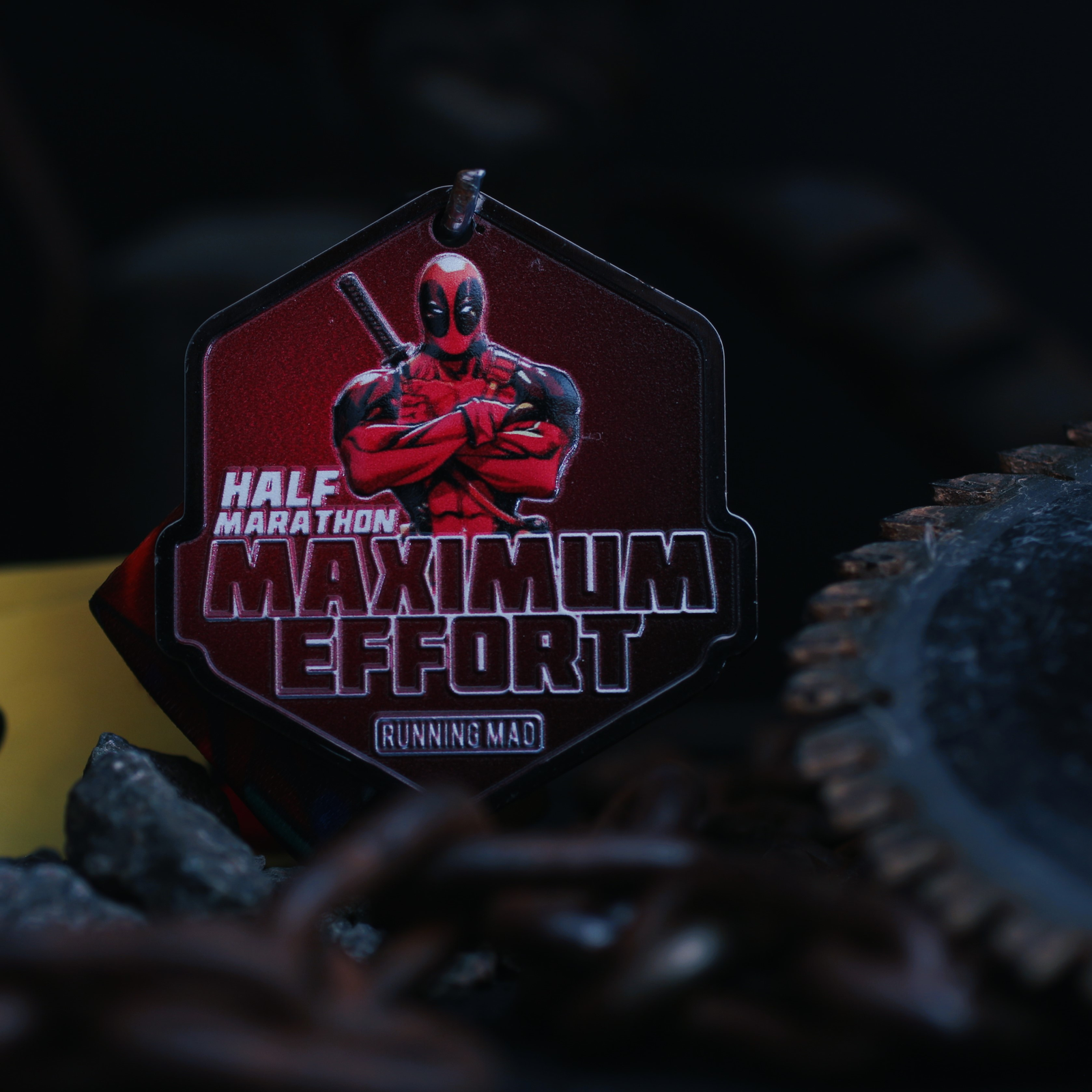 A medal with an illustration of a red-suited character holding swords, with text reading "Half Marathon Maximum Effort" and "Running Mad." The medal hangs among chains and gears, set against a dark background.
