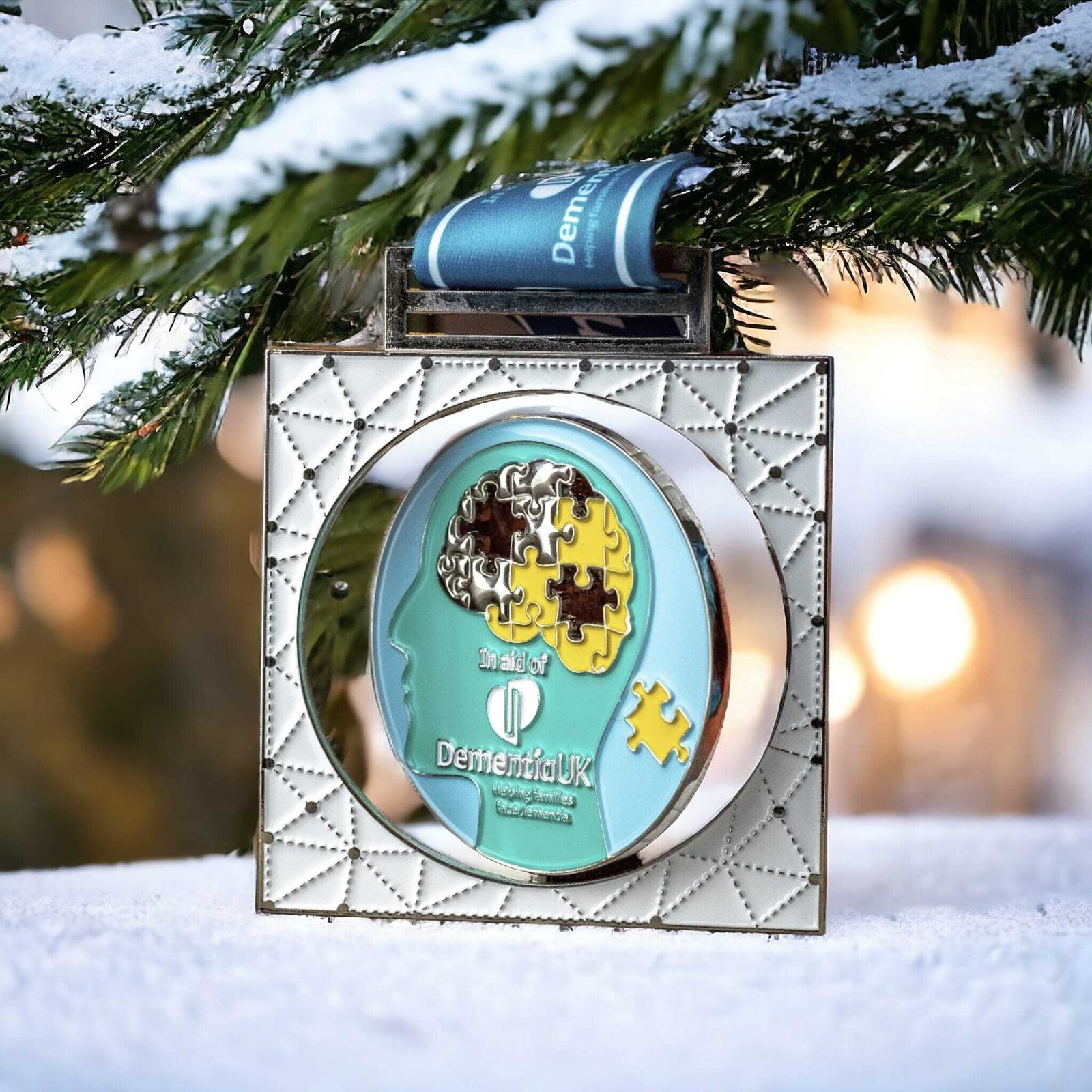 A medal with a puzzle piece design depicting a human head is hanging on a snow-covered branch. The medal features the text "in aid of Dementia UK" and has a blue ribbon.