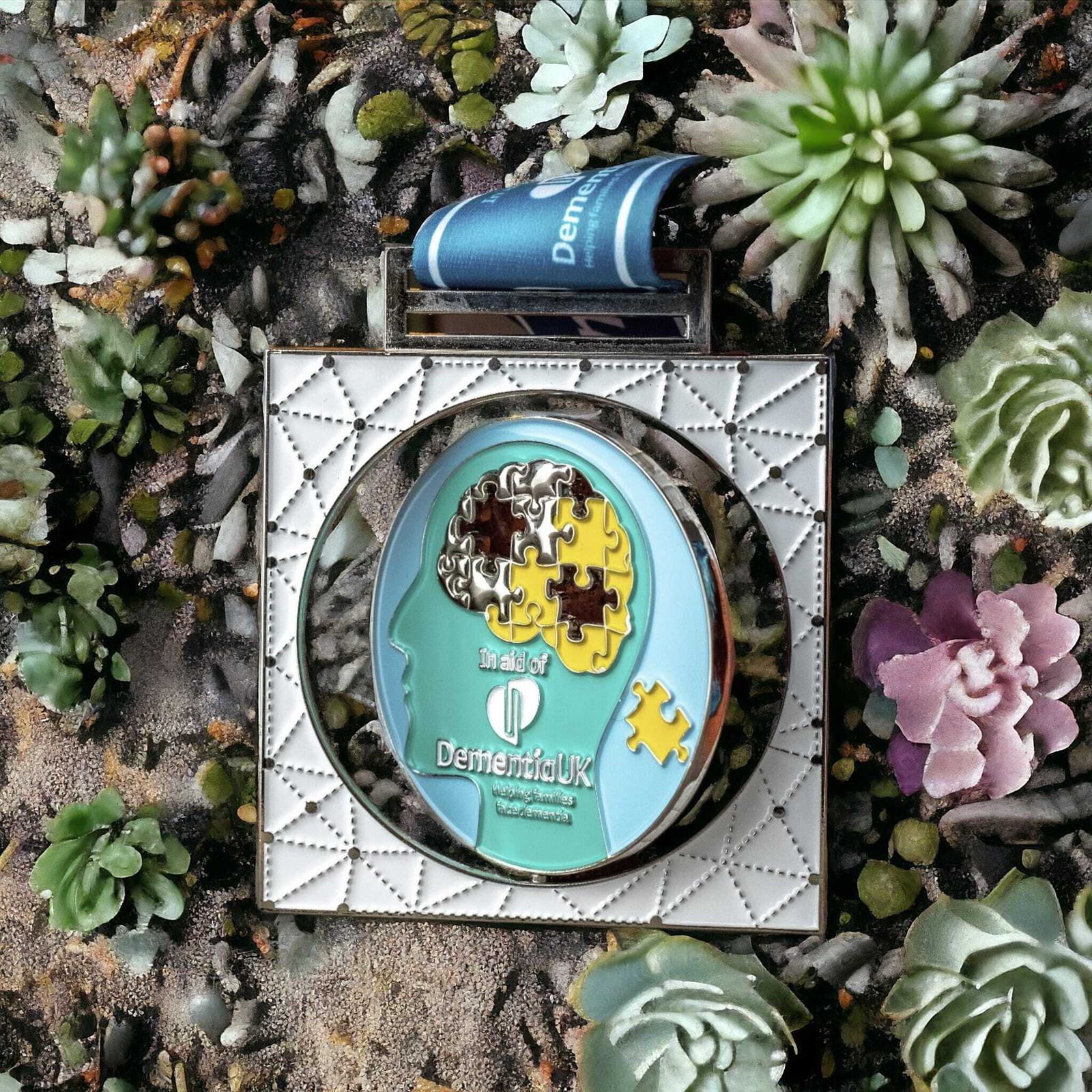 A medal with a puzzle piece design incorporating a human head and Dementia UK logo is surrounded by a border resembling a perfume bottle, set amidst assorted succulents.