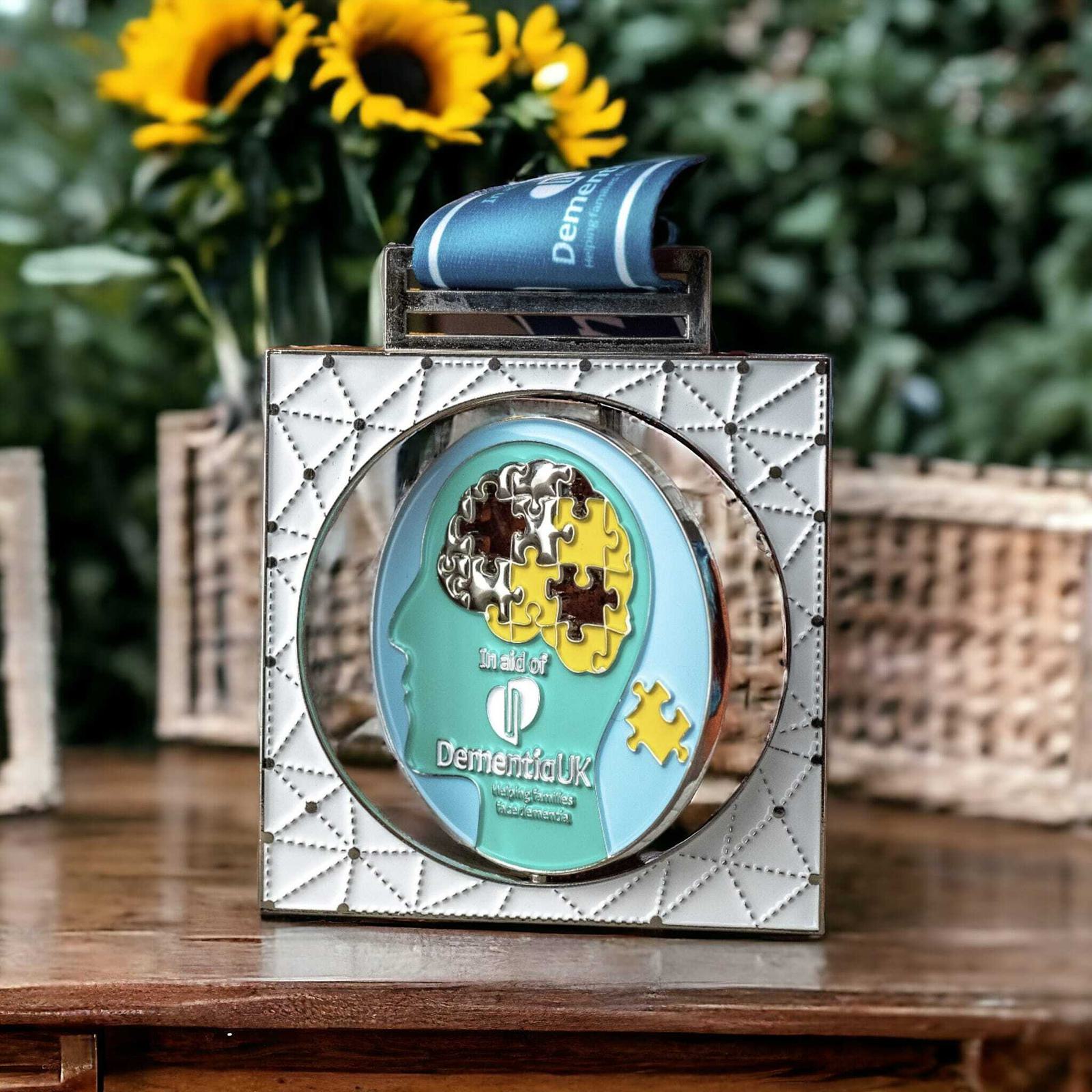 A medal sits in a decorative frame on a wooden table, with puzzle pieces forming a human brain on its surface. The text reads "Dementia UK." Sunflowers and greenery are blurred in the background.