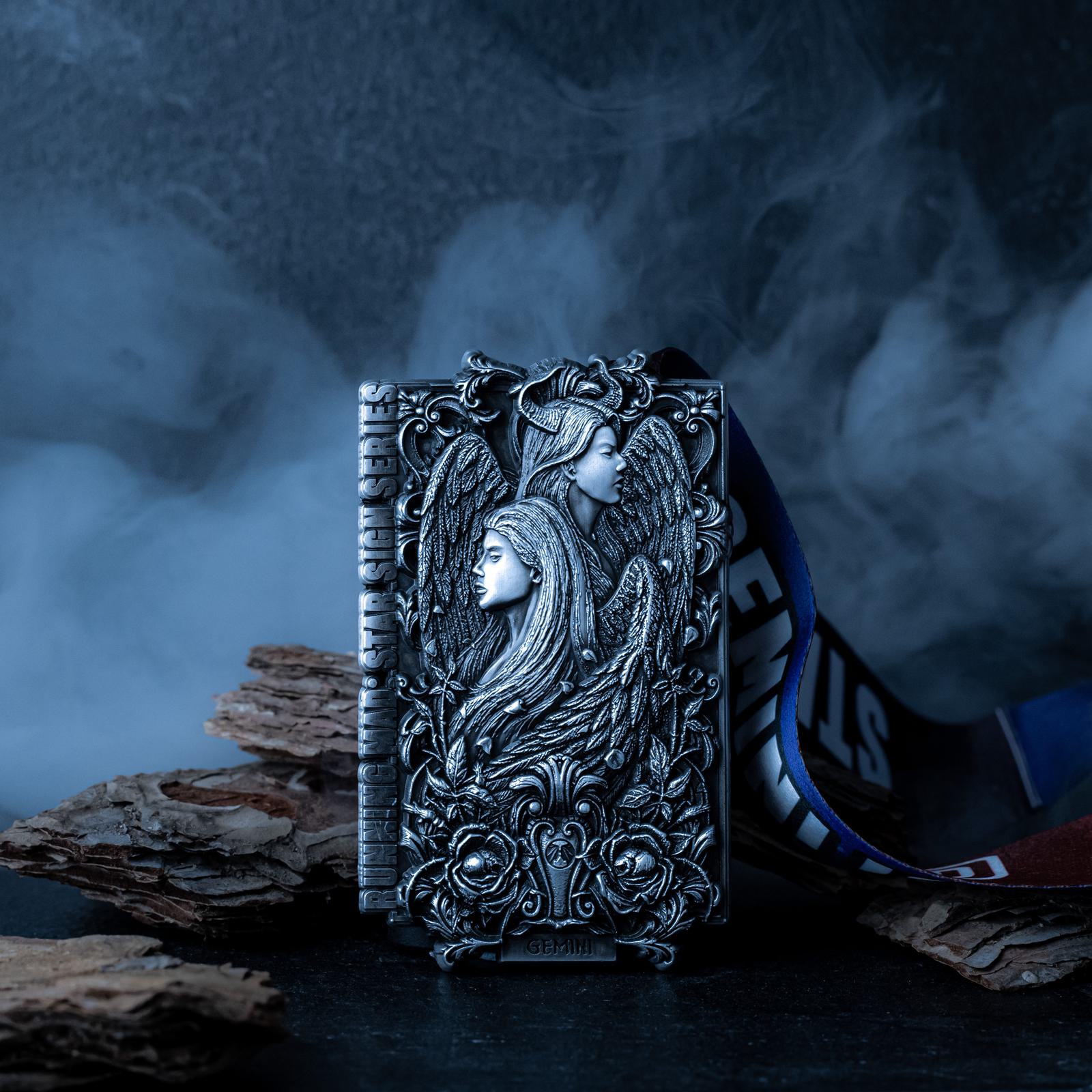 A silver rectangular object with intricate engravings of winged figures and floral patterns stands against a smoky background. Pieces of bark and a colorful ribbon are in the backdrop, creating a mystical atmosphere.