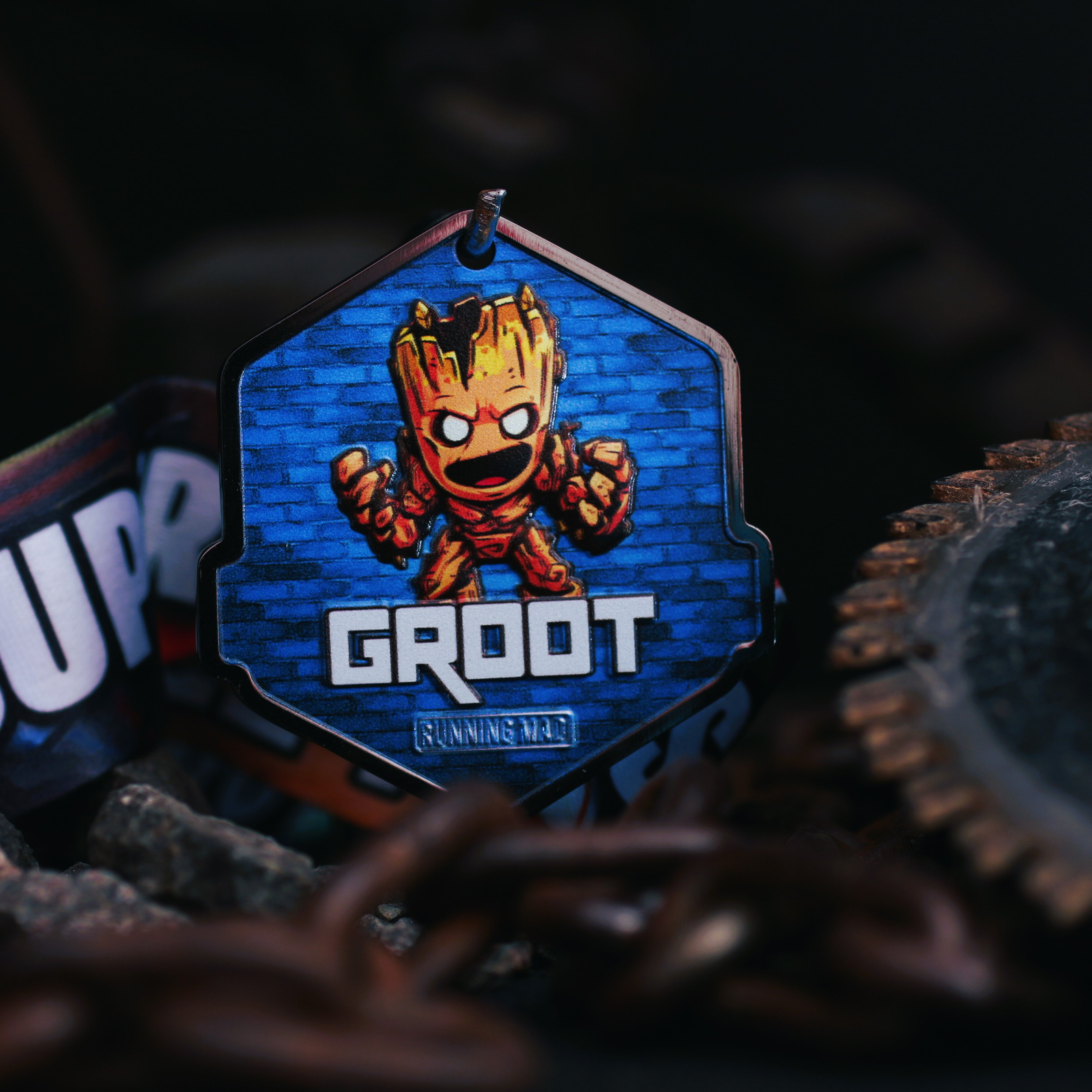 A keychain featuring a cartoonish figure resembling a small tree with a smile, labeled "Groot." It has a blue background with rustic chains and gears in the foreground, giving an industrial vibe.