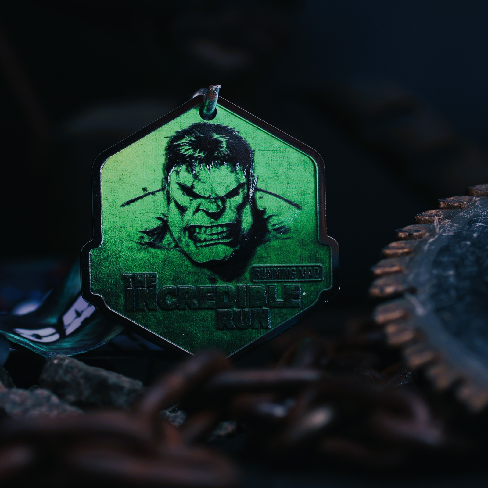 A close-up of a medal featuring a green illustration of an angry, muscular figure with the text "The Incredible Run" underneath. Chains and industrial gears surround the medal against a dark background.