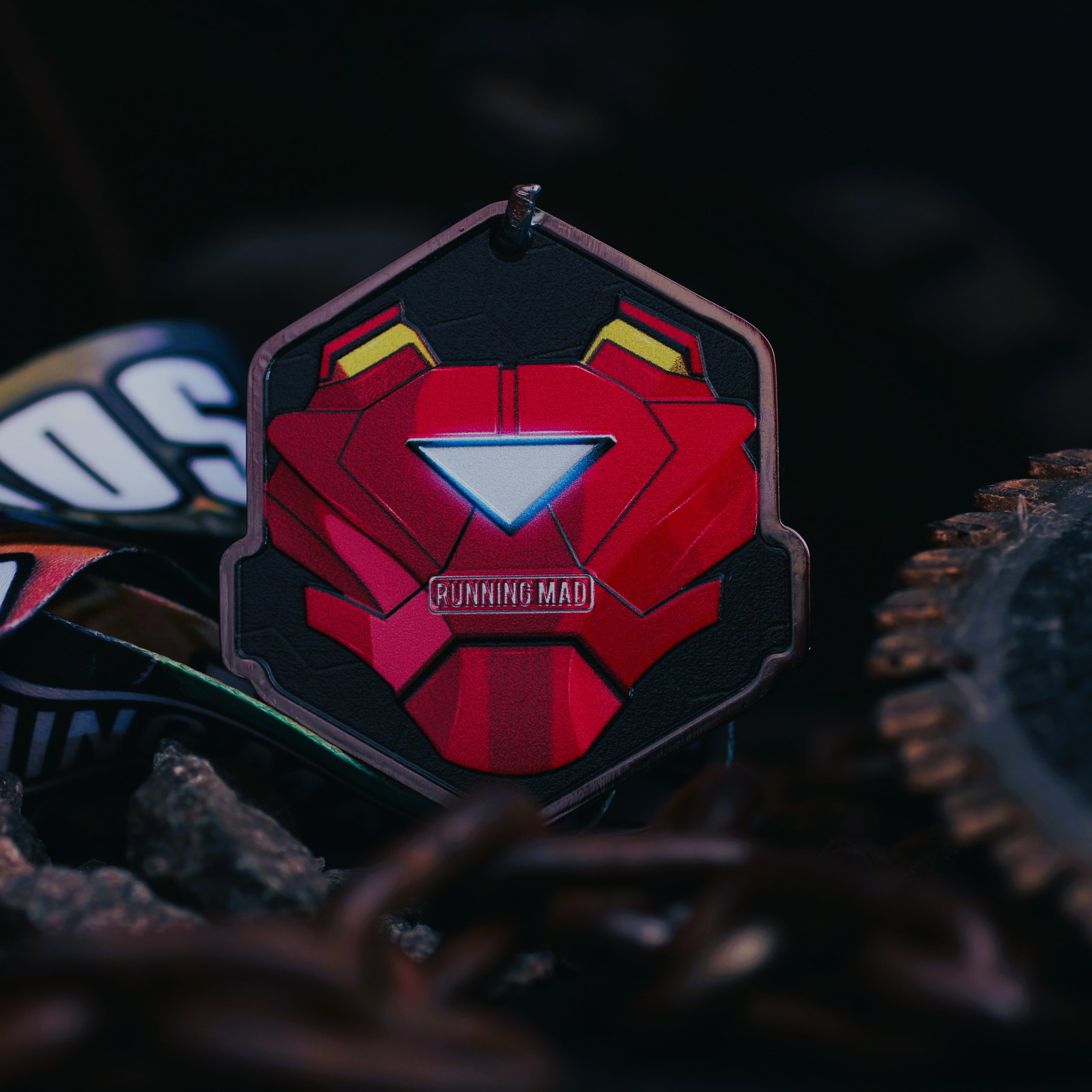 A medal in the shape of a red mask with a triangular white light in the center is surrounded by a dark background. The text "RUNNING MAD" is inscribed beneath the light. Chains and blurred objects are visible around the medal.