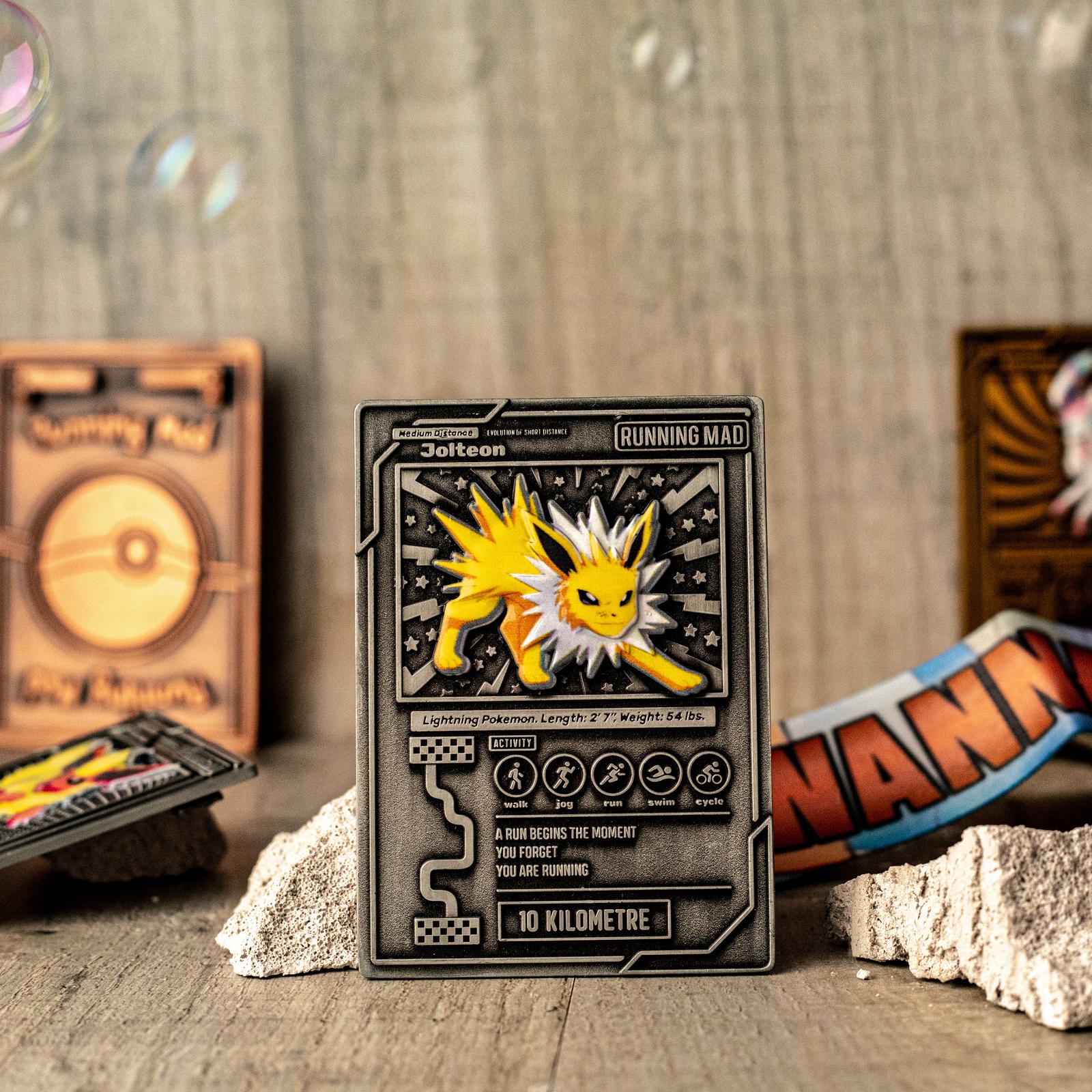 A metal card featuring Jolteon, a Pokémon, displayed on a rock. The card includes detailed stats and the phrase "Running Mad." In the background are other Pokémon-themed items and a colorful NAM open band on rocks.