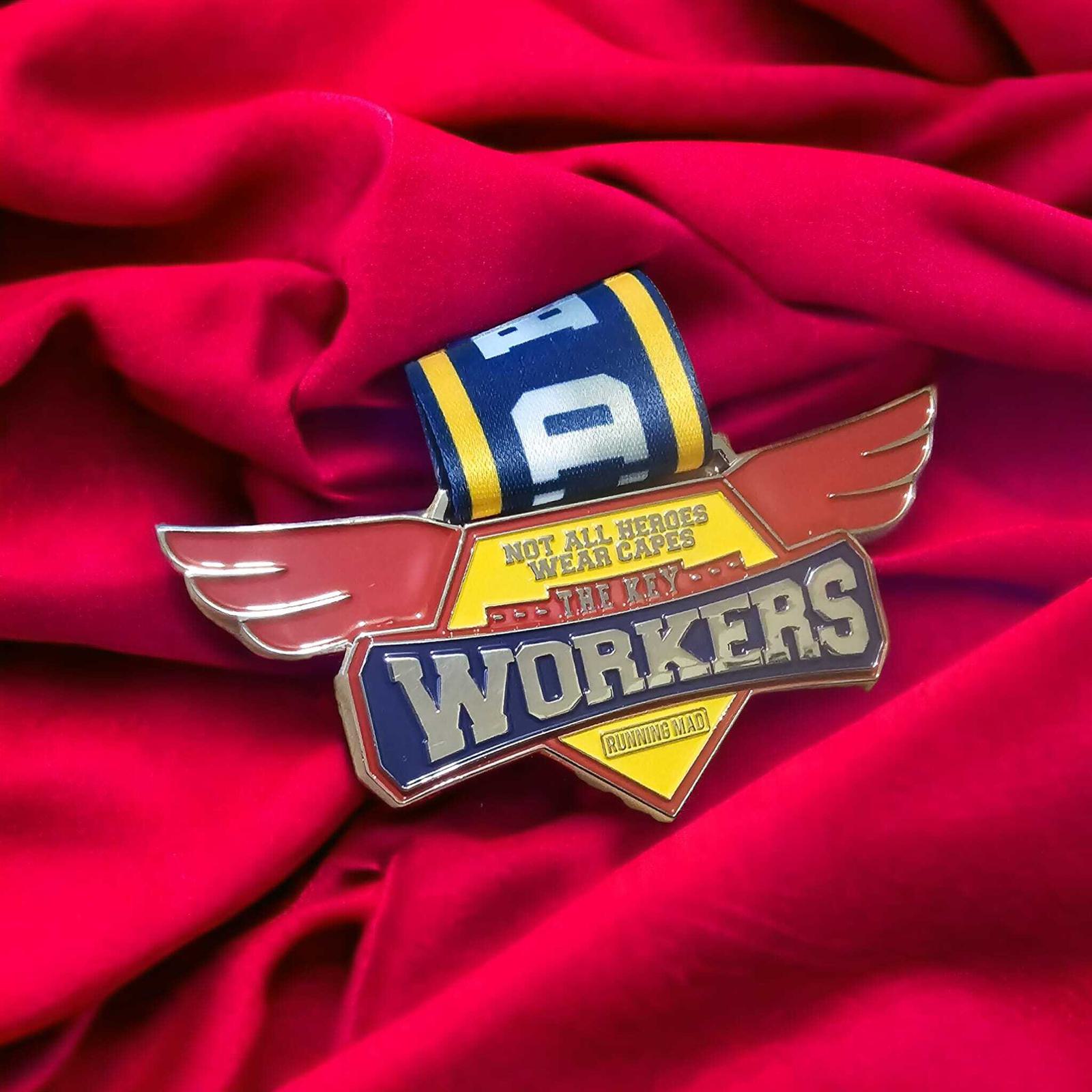 A medal with a winged design and the words "Not all heroes wear capes, they are workers" lies on red fabric. The top has a blue and yellow ribbon with numbers.