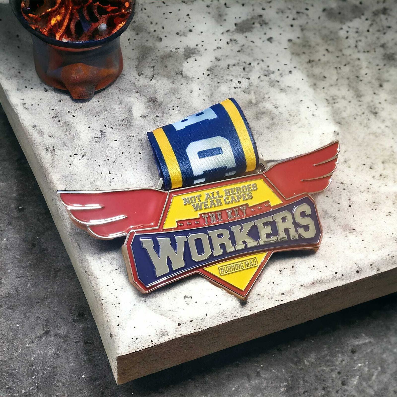 A colorful badge with wings reads "Not All Heroes Wear Capes, In This Family Workers Build the Road." It's placed on a concrete surface next to a piece of rolled-up, blue and yellow striped cloth.