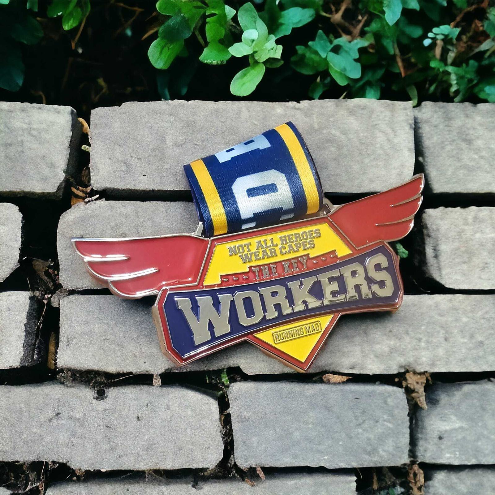A badge resting on stone bricks, featuring red wings and text: "Not all heroes wear capes, they are workers." A yellow, blue, and white ribbon is attached above it. Green leaves are in the background.